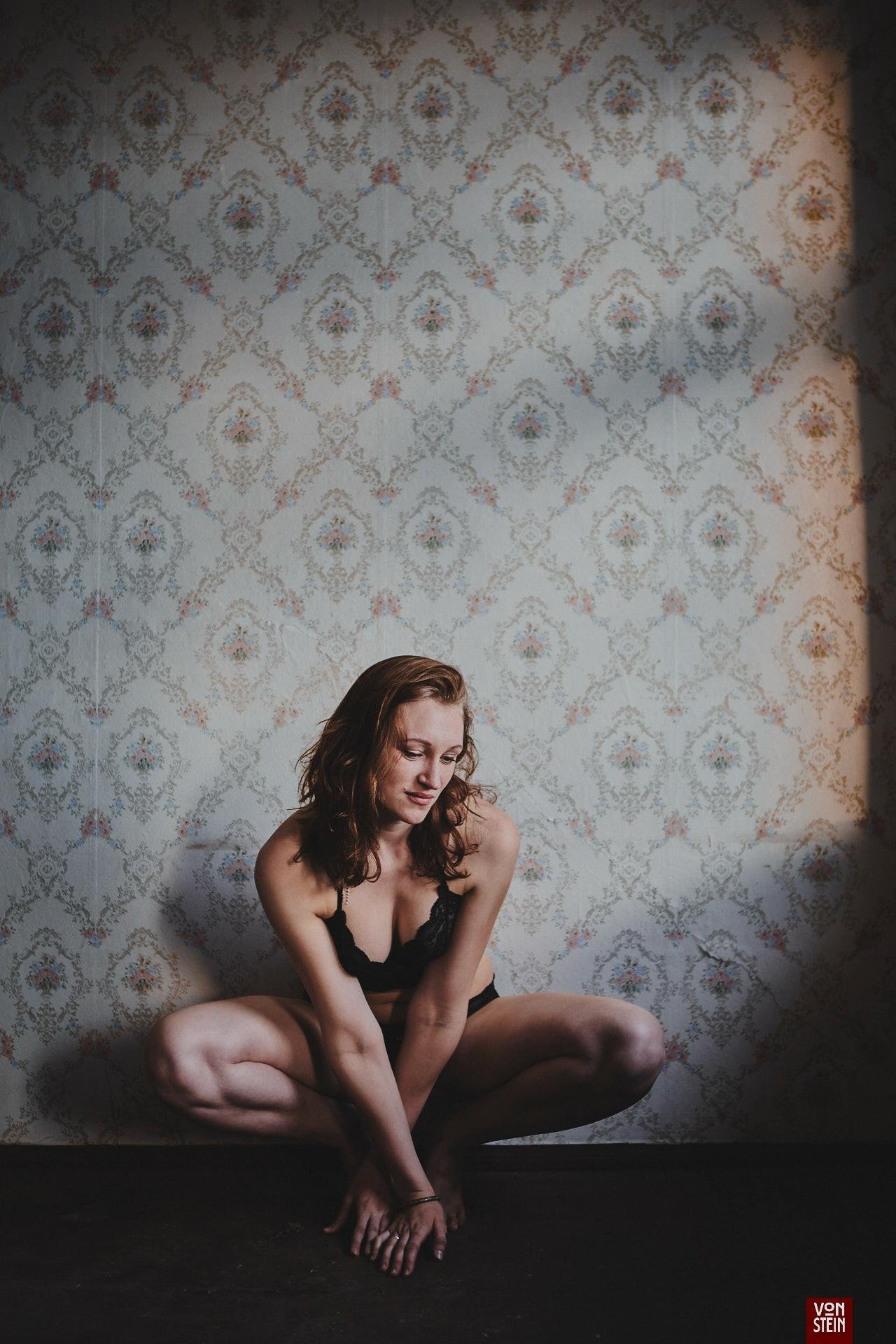 Woman in black lingerie crouching on the floor with knees apart and hands on the ground, looking down, against a pale patterned wallpaper background with vignette darkening around the edges.