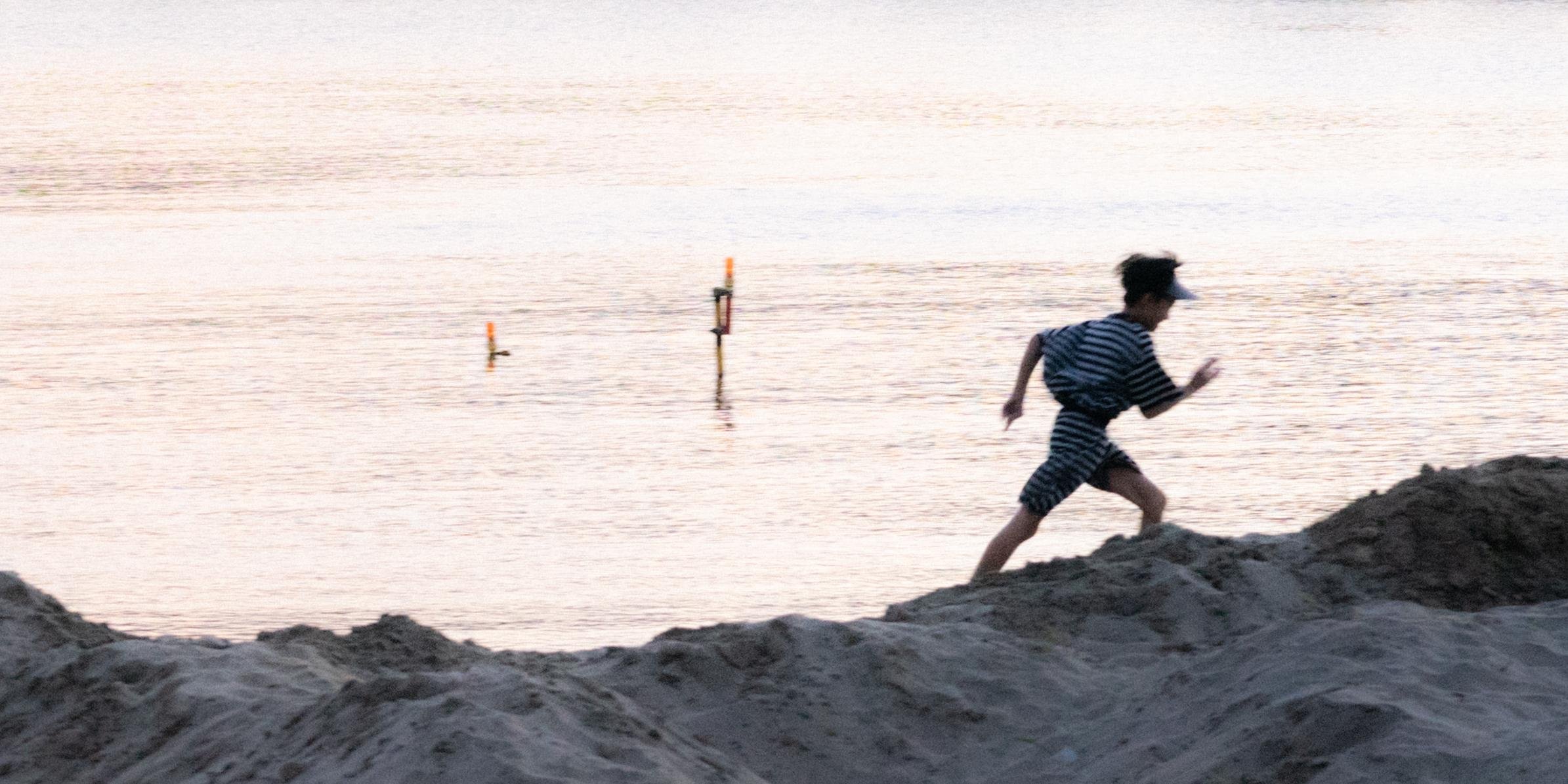 A person wearing a striped shirt and shorts runs along a sandy beach. The background features calm water with two orange markers partially visible above the surface. The sky and water have a soft, pastel hue, suggesting early morning or late afternoon lighting.