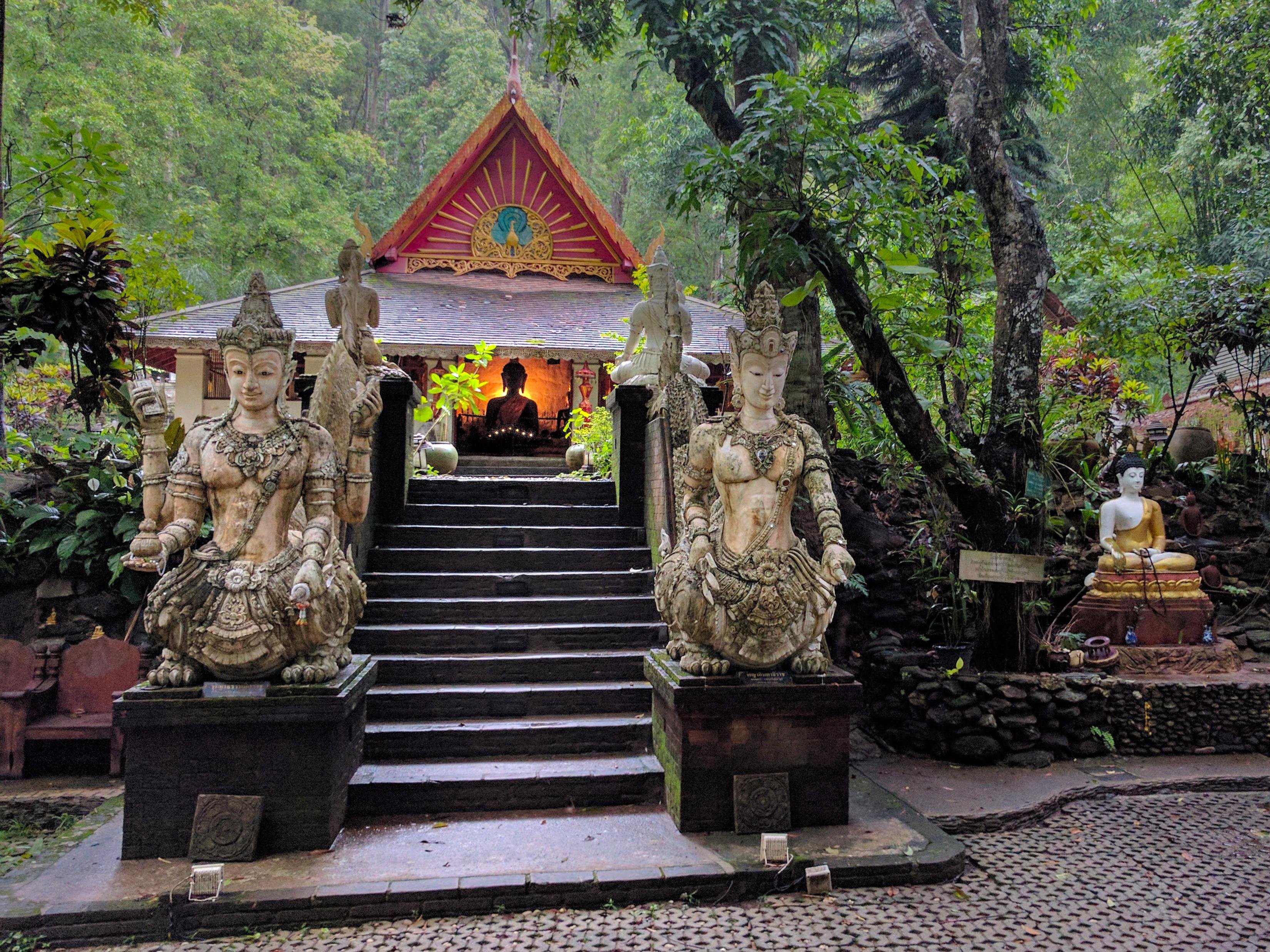 Stone staircase leading to a small temple pavilion with a red gabled roof, flanked by two large seated guardian statues on pedestals; dense green forest surrounds the site, with a Buddha figure visible in the lit interior and another seated statue to the right.