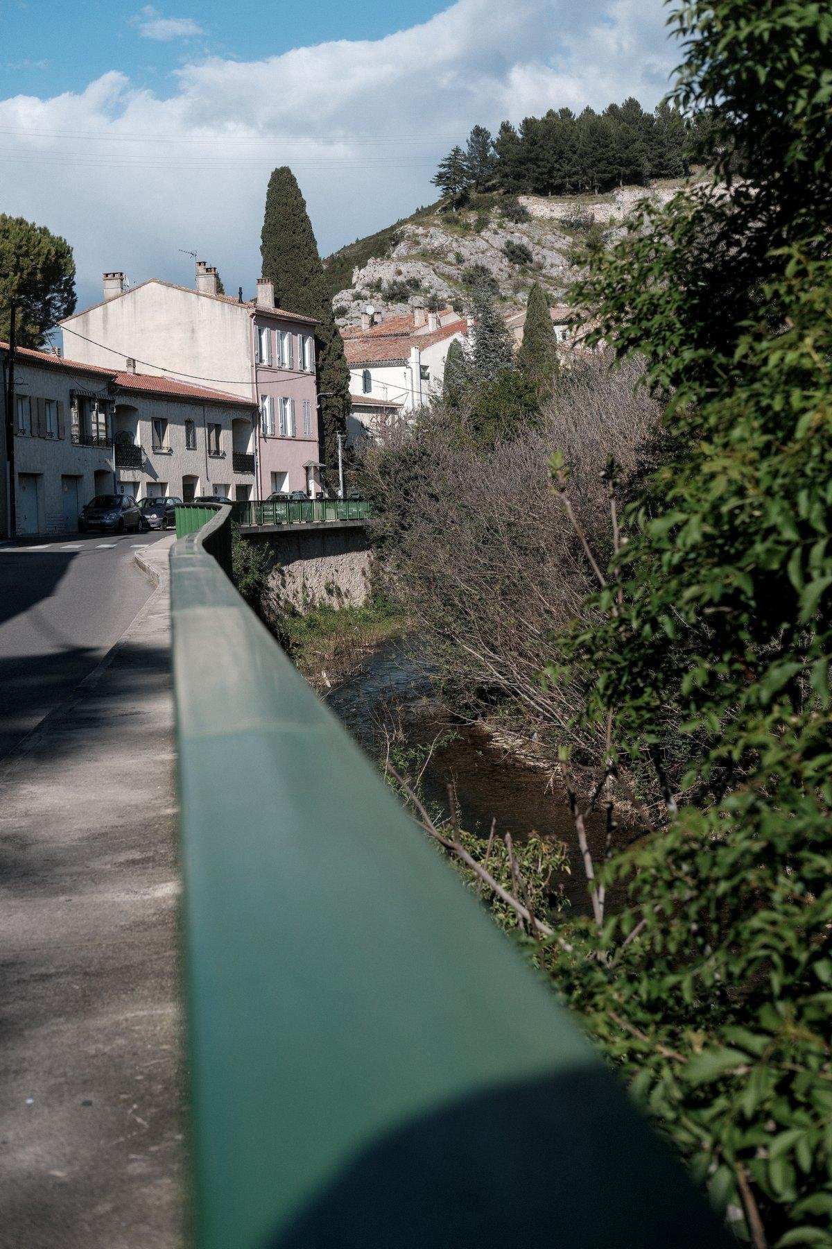 Photo verticale. Vue à ras d’une rambarde verte de sécurité qui part au loin, avec à gauche le trottoir et la route et à droite, une rivière. Au fond des habitations de faible hauteur et une montagne