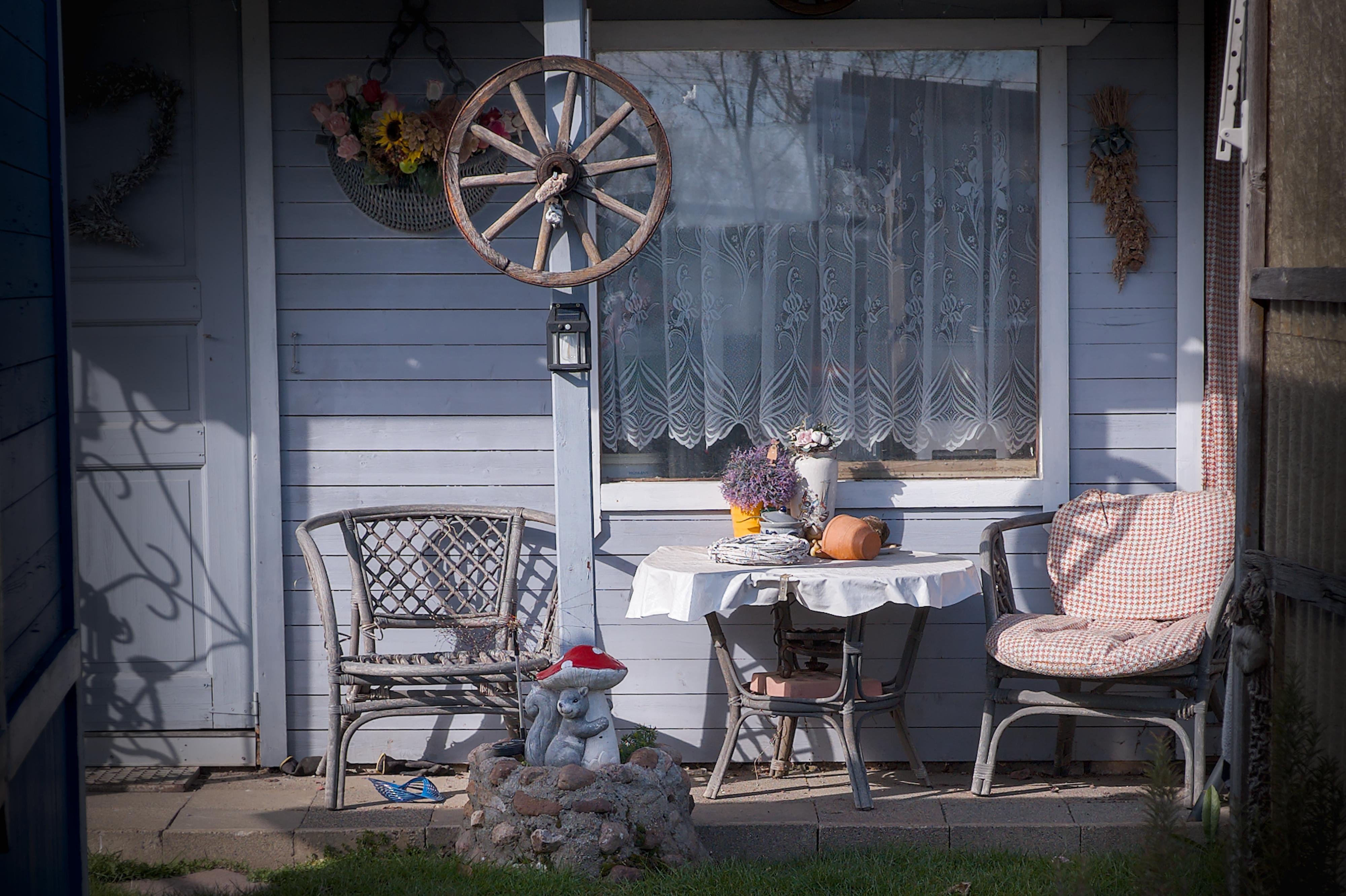 Small patio against a light blue wooden wall with a lace-curtained window; two wicker chairs flank a round table with a white cloth holding a vase of flowers, a purple plant, and a pumpkin, with a wagon wheel decoration above and a small garden statue wearing a red hat in the foreground.
###
Kleine Terrasse gegen eine hellblaue Holzwand mit einem Spitzenfenster; zwei Korbstühle flankieren einen runden Tisch mit einem weißen Tuch mit einer Vase Blumen, einer lila Pflanze und einem Kürbis, mit einer Wagenraddekoration darüber und einer kleinen Gartenstatue mit einem roten Hut im Vordergrund.