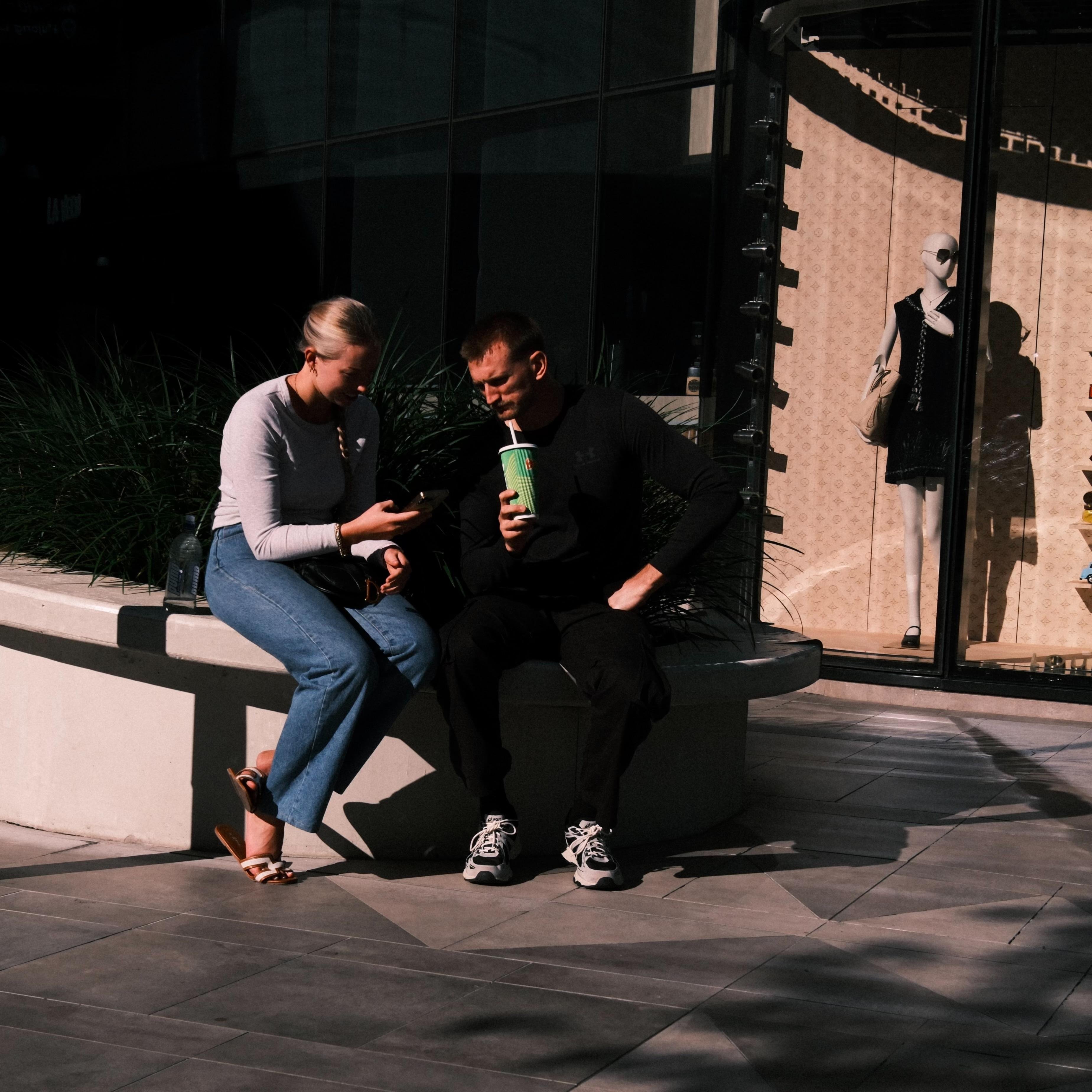 Two people sit on a concrete planter outside a storefront; the woman looks at a phone while the man holds a green drink cup, with ornamental grasses behind them and a mannequin in the shop window on the right.