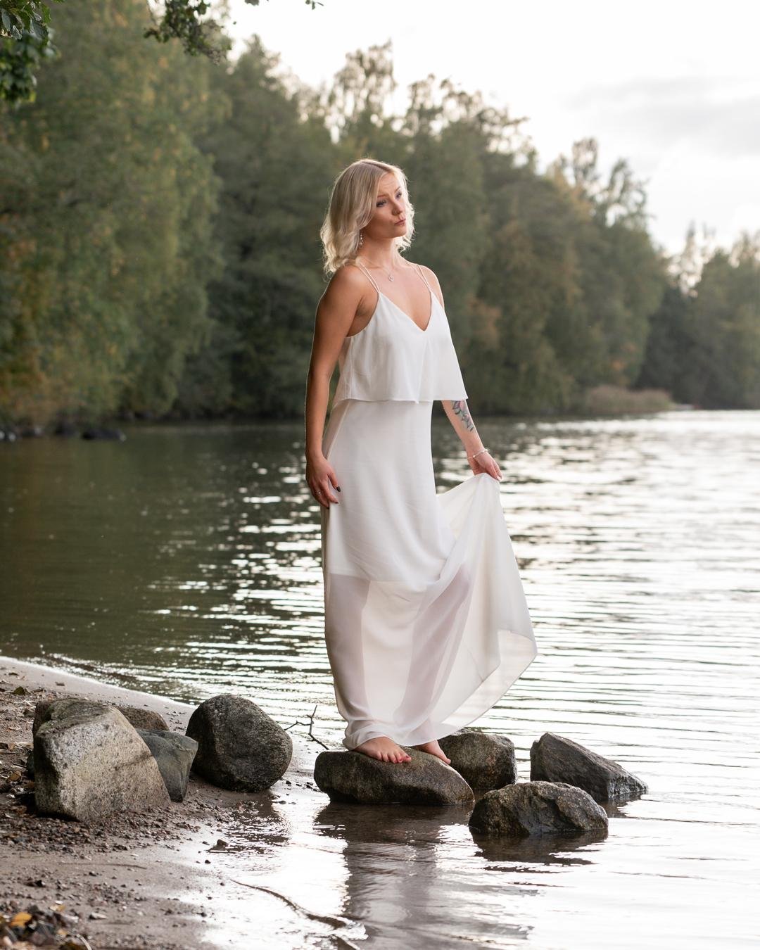 The image shows a person standing on rocks by the edge of a body of water. They are wearing a long, white dress, and there are trees in the background. It appears to be a natural, outdoor setting.