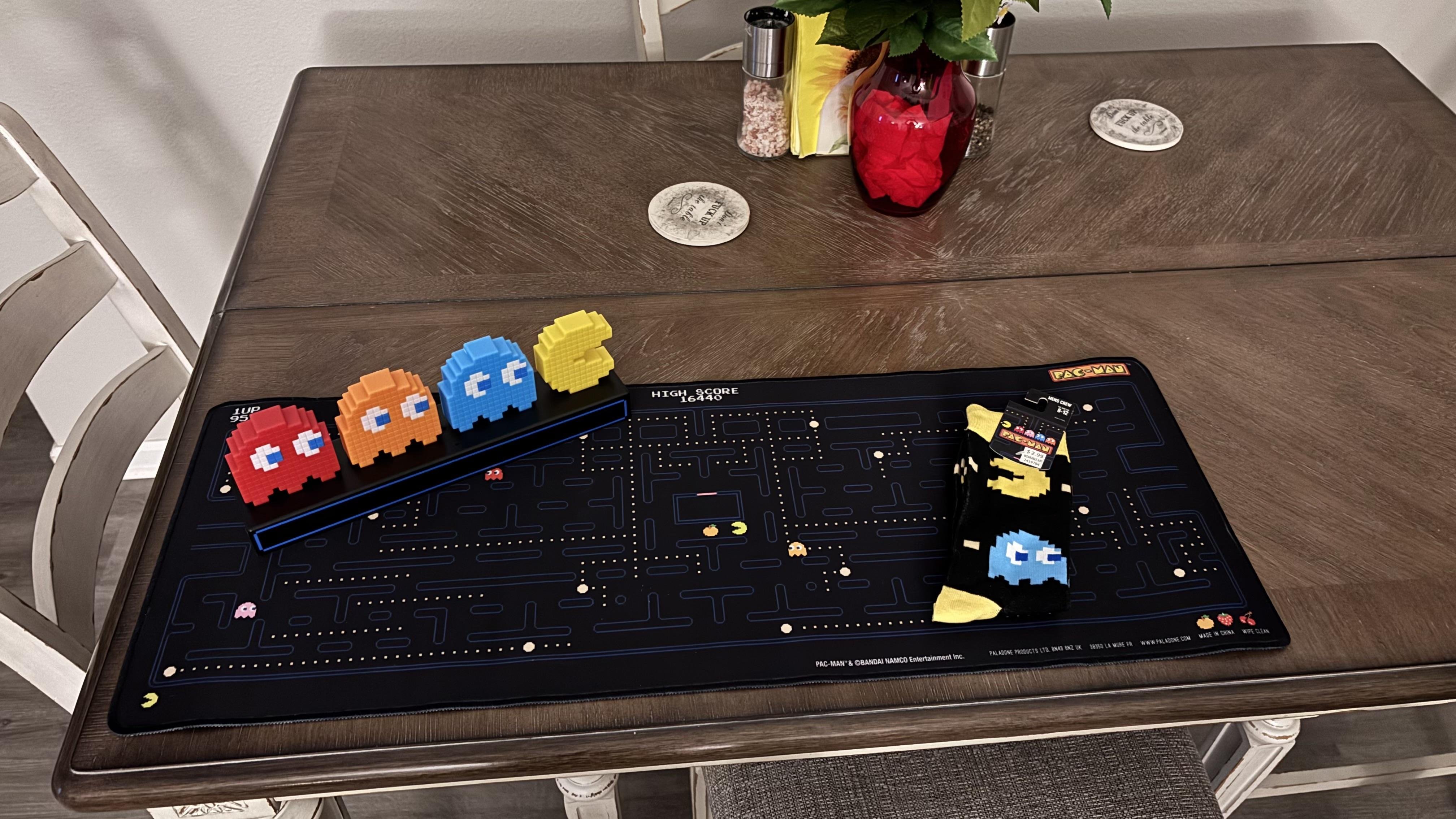 A high-angle shot depicts a dark wood table covered in Pac-Man memorabilia. On the left side of the table is a row of three-dimensional pixelated Pac-Man characters in red, orange, and blue, followed by a Pac-Man figure. These characters are arranged on a black, rectangular stand. The table is covered by a black Pac-Man game-themed mat, with a maze design and small Pac-Man and ghost figures scattered throughout. A pair of black Pac-Man socks sits on the right side of the mat. In the background, there is a vase with red flowers and greenery, along with two small glass containers. A decorative coaster is also visible on the table's surface. A white chair is situated on the left side of the table.