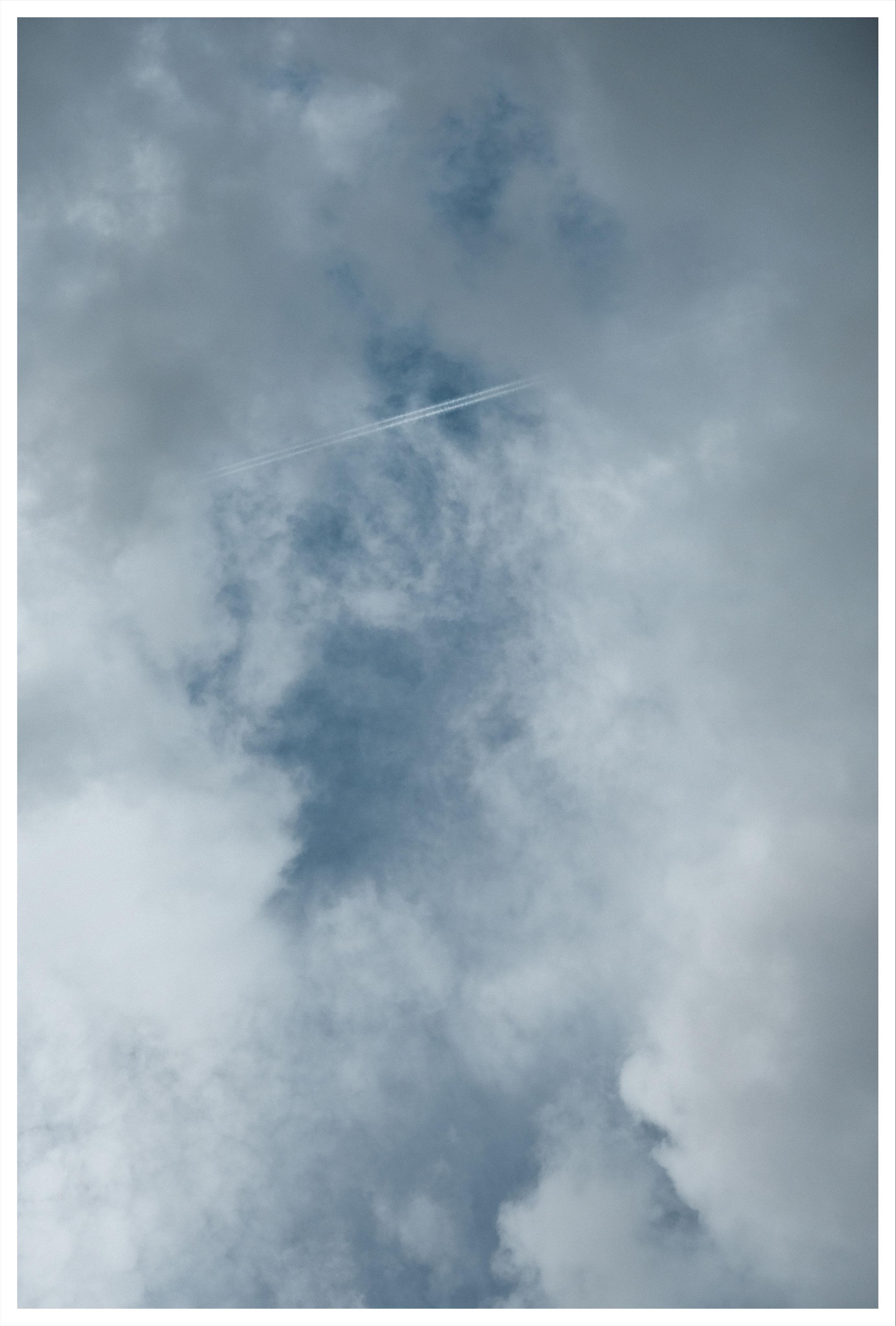 View straight up onto the cloudy sky. In an opening in the thick clouds a straight contrail is visible on the blue sky.