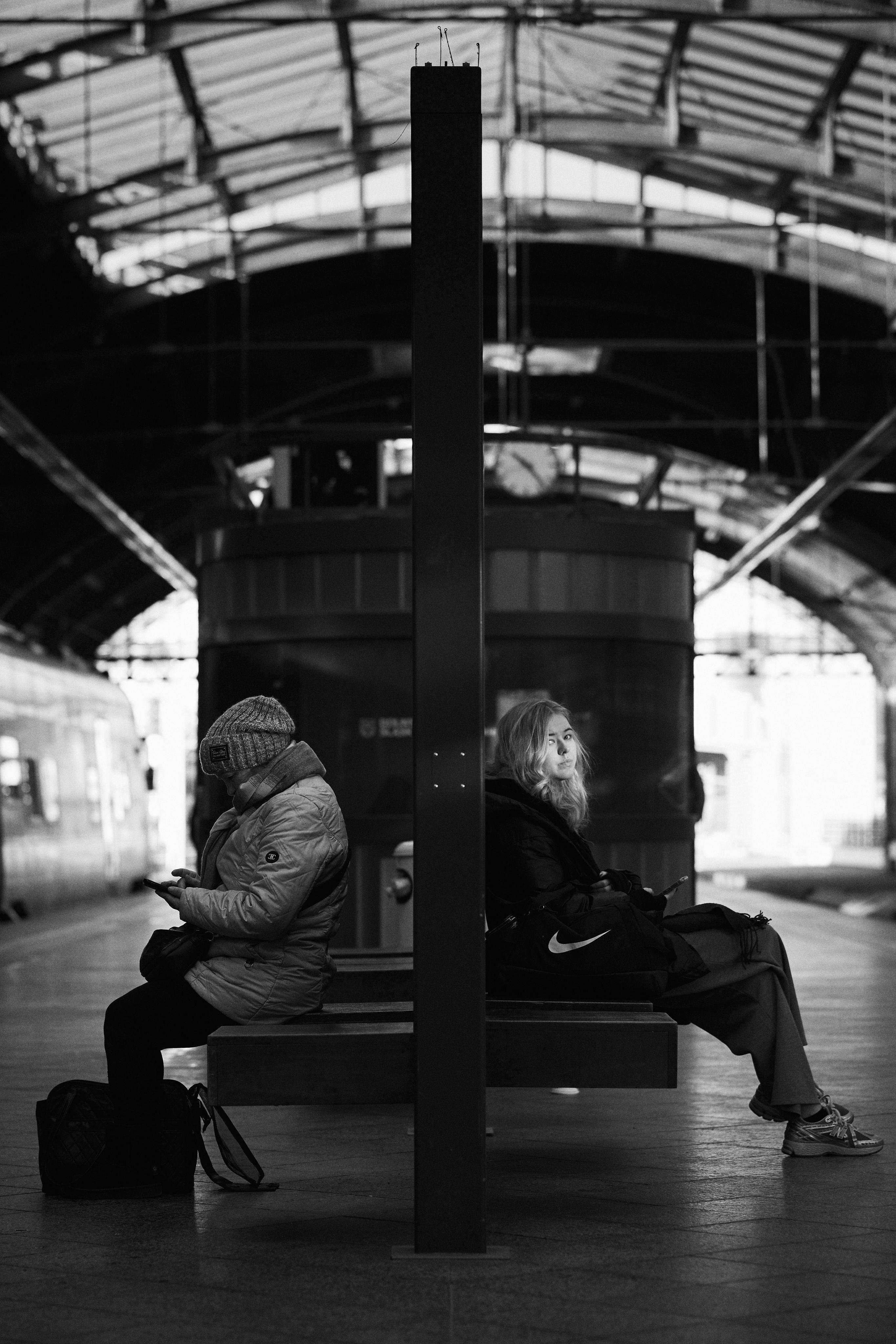 The image depicts a black and white scene at a train station. Two people are sitting back to back on a bench. One person is wearing a beanie and looking at a phone, while the other person is looking forward, sitting with luggage next to them. The station has a large arched ceiling and a train is visible on the left side.