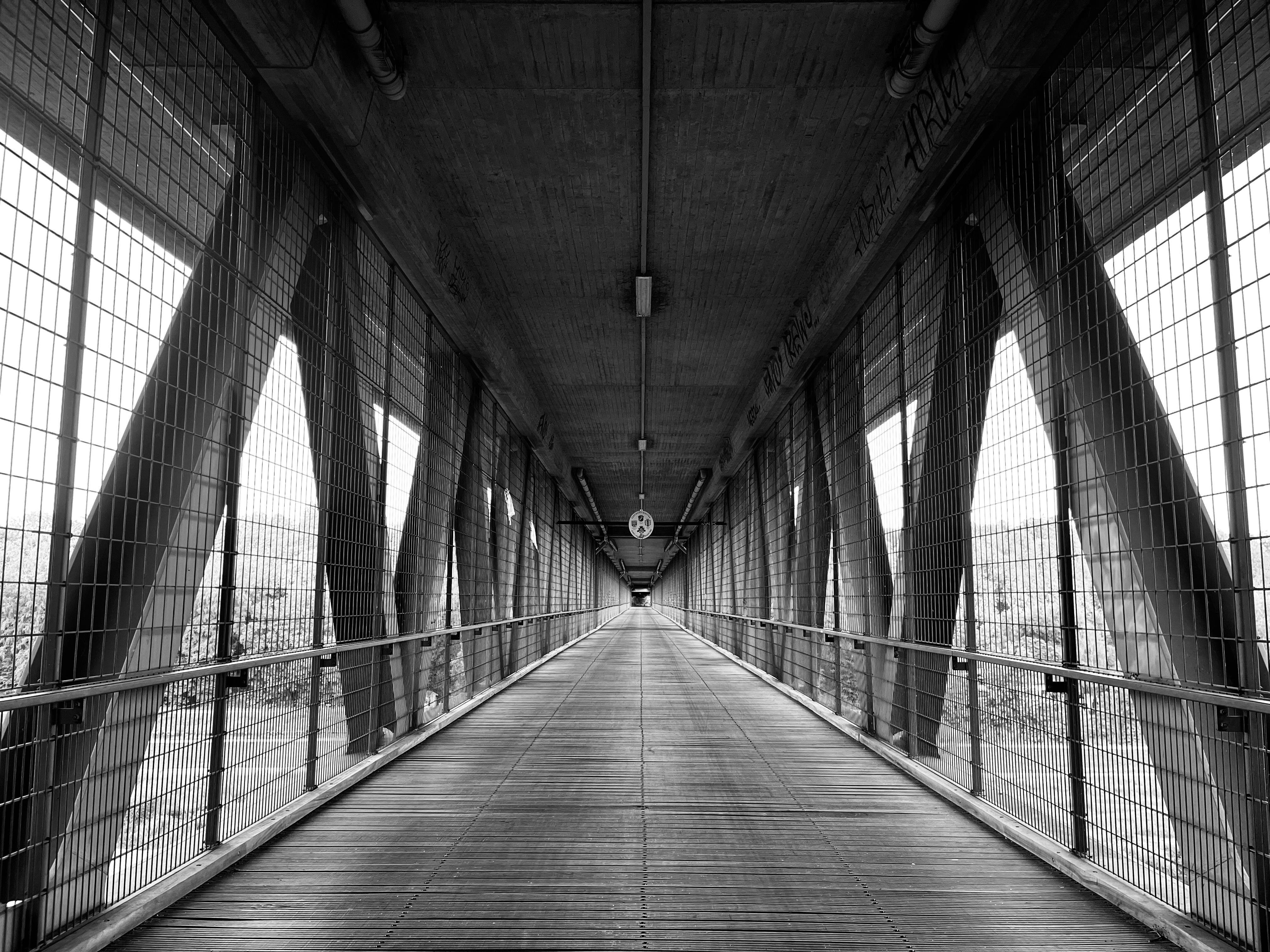 Black-and-white view down a long enclosed pedestrian truss bridge with wooden plank flooring, wire-mesh side walls, metal railings, and diagonal steel supports, converging toward a central vanishing point under a concrete ceiling with pipes and lights.