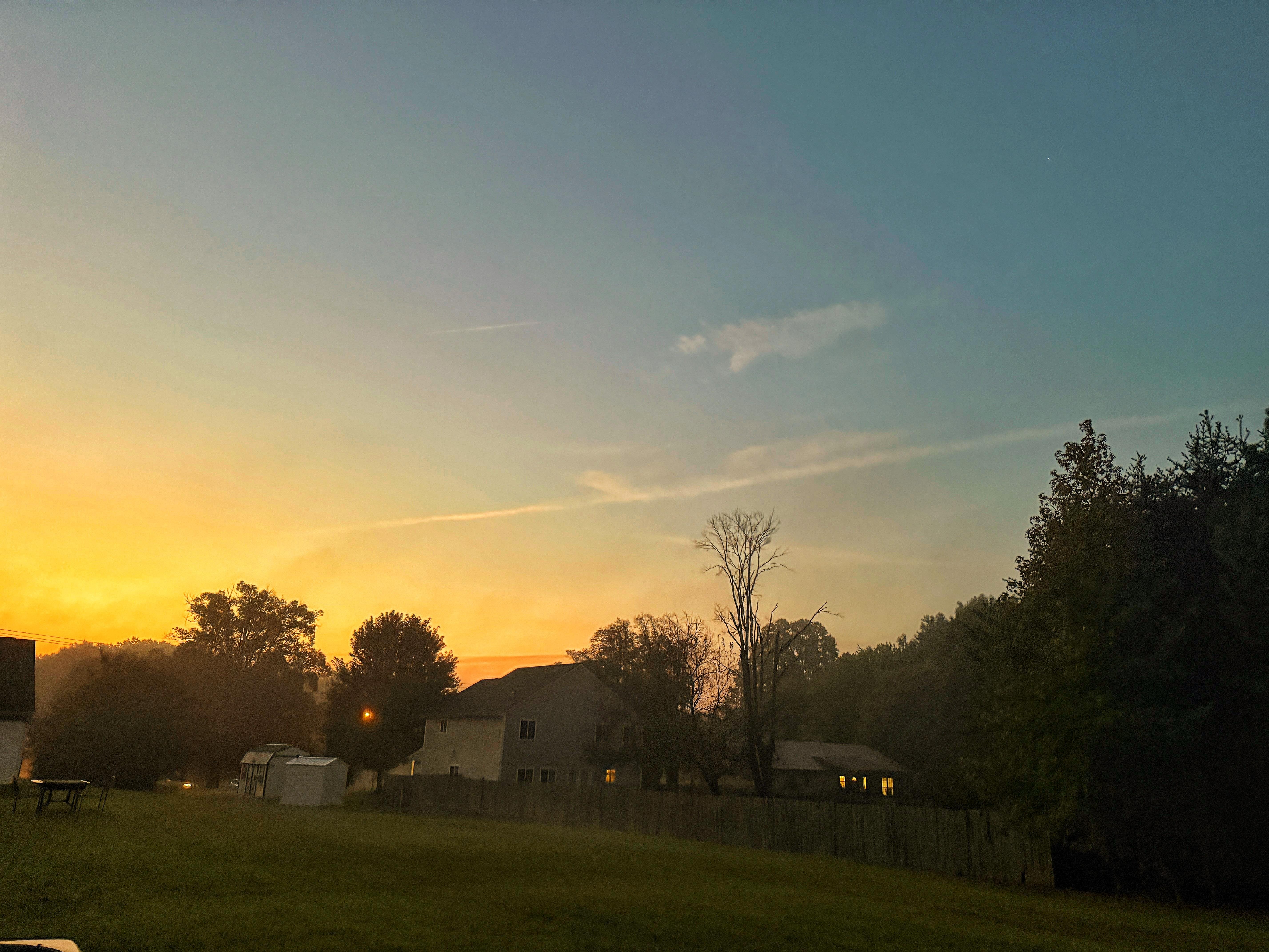 The image depicts a serene suburban scene at sunset. The sky is a gradient of warm colors, transitioning from a deep blue at the top to a vibrant orange near the horizon, where the sun is setting. Wispy clouds are scattered across the sky, illuminated by the setting sun. In the foreground, there is a grassy field with a wooden fence running along the right side, separating the field from a line of trees. Several houses are visible in the background, with one having a lit window, suggesting activity inside. A small shed is also present in the scene. The overall atmosphere is calm and peaceful, with the warm colors of the sunset casting a soft glow over the landscape.