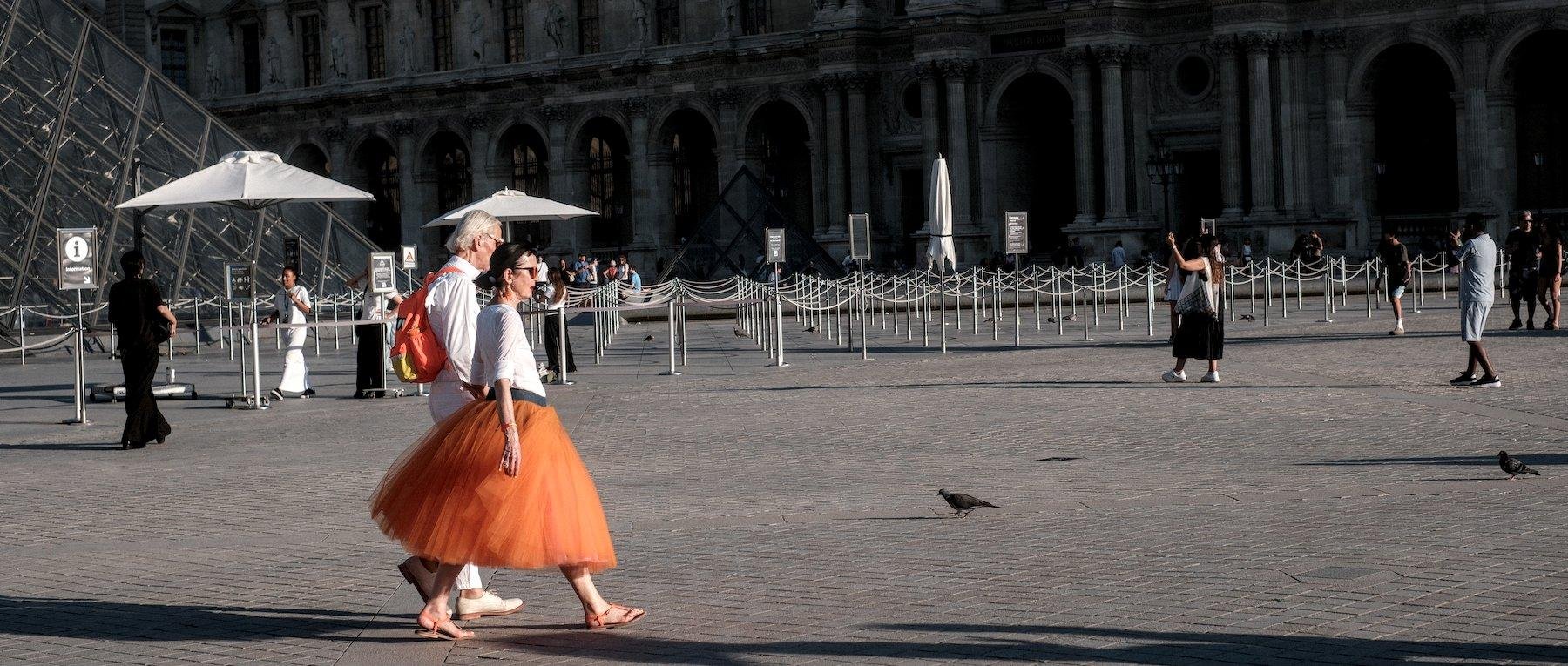 Photo horizontale au ratio cinemascope. Un couple de personnes âgées marche sur la place du Louvre. La femme porte une longue jupe bouffante en tulle orange contrastant avec le gris des pavés