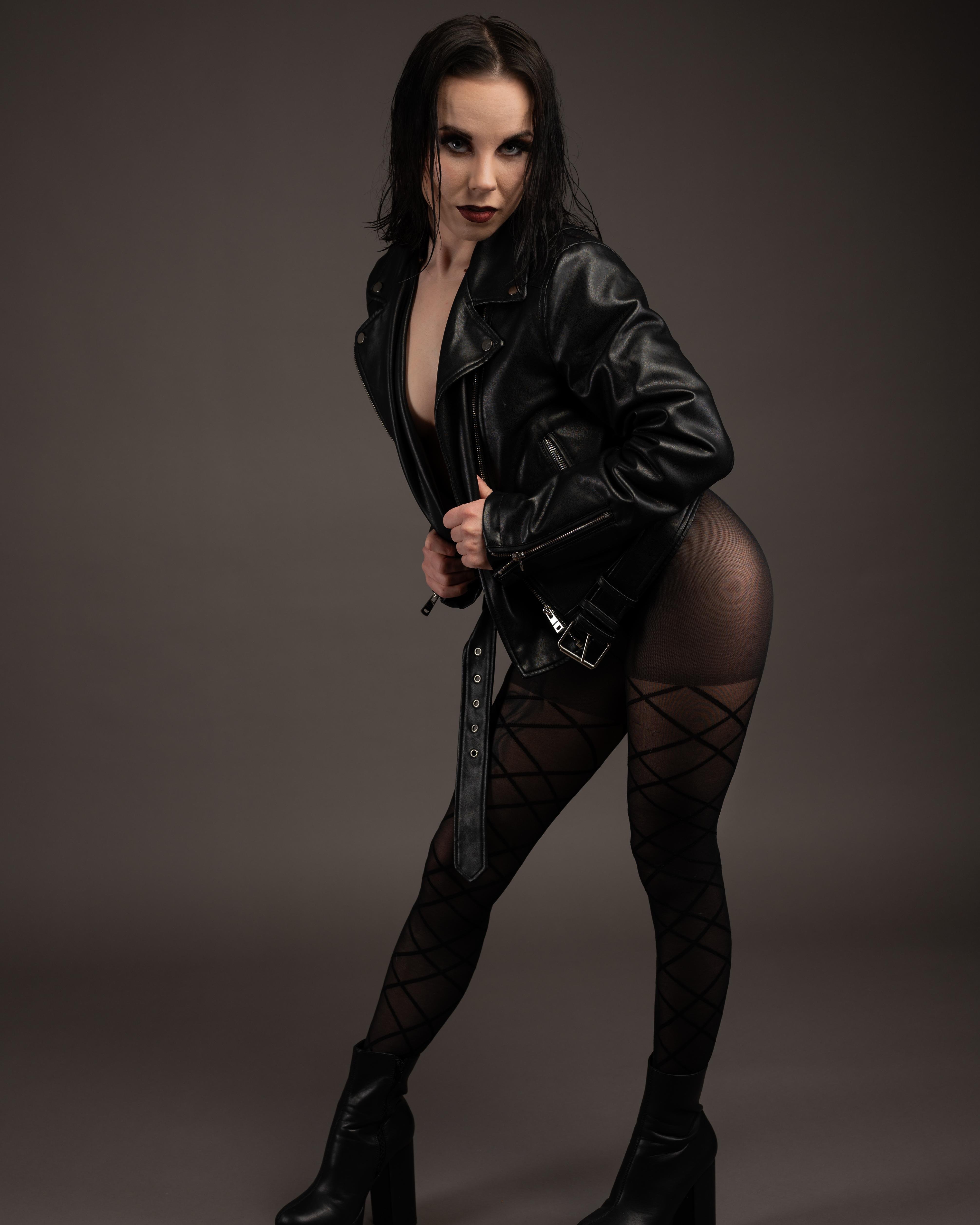 Woman with dark hair wearing a black leather jacket over a dark outfit, posing against a plain gray background. Her tights feature a crisscross pattern, and she wears high platform boots.