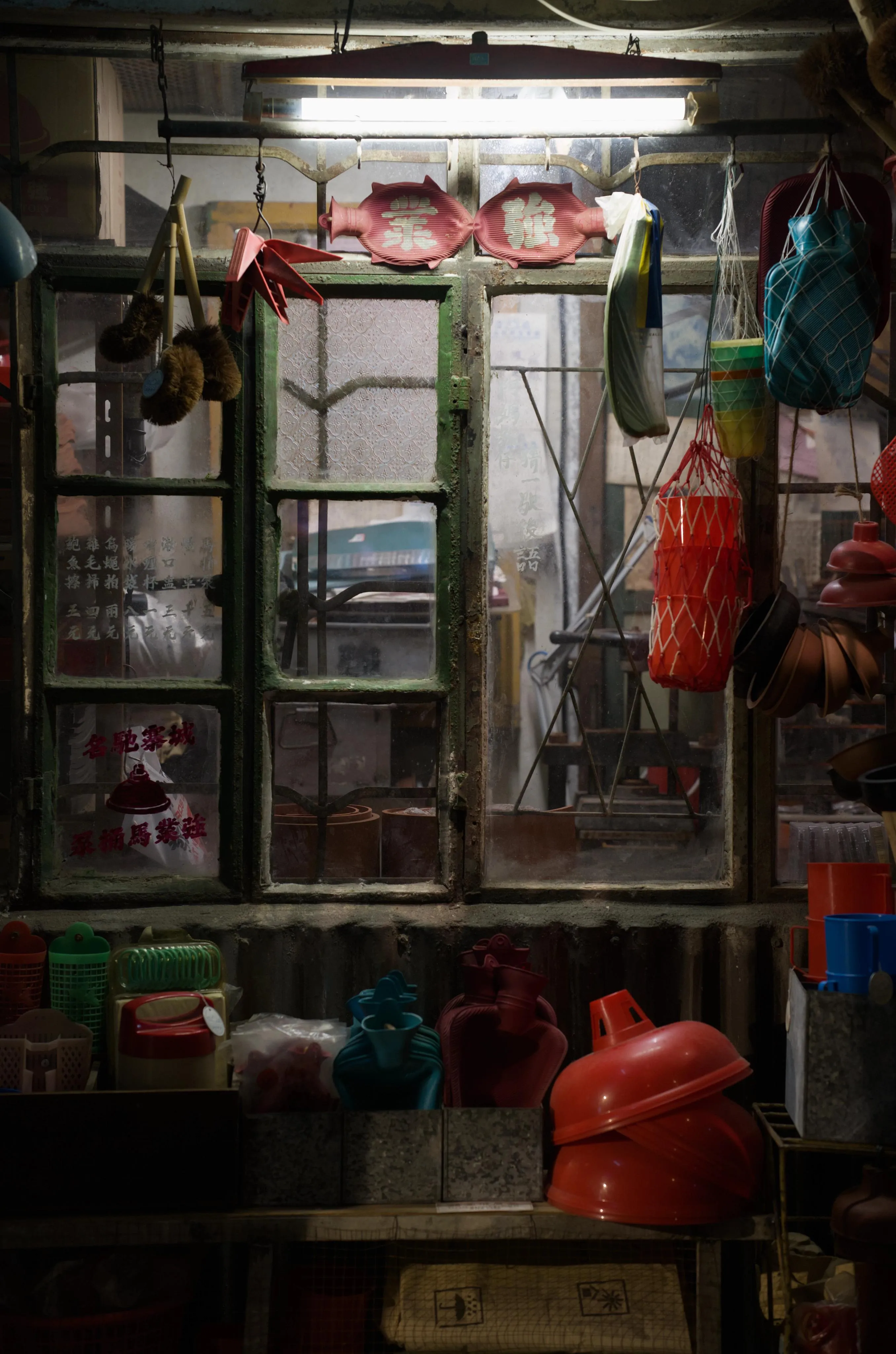 Photograph of a window between two indoor rooms with various household objects hanging under, over and around the window. Most of the household objects are plastic and have bright colors such as red, blue and green. The window appears to be weathered, rusty green metal. A neon bar-shaped white light lights the window from its top.
