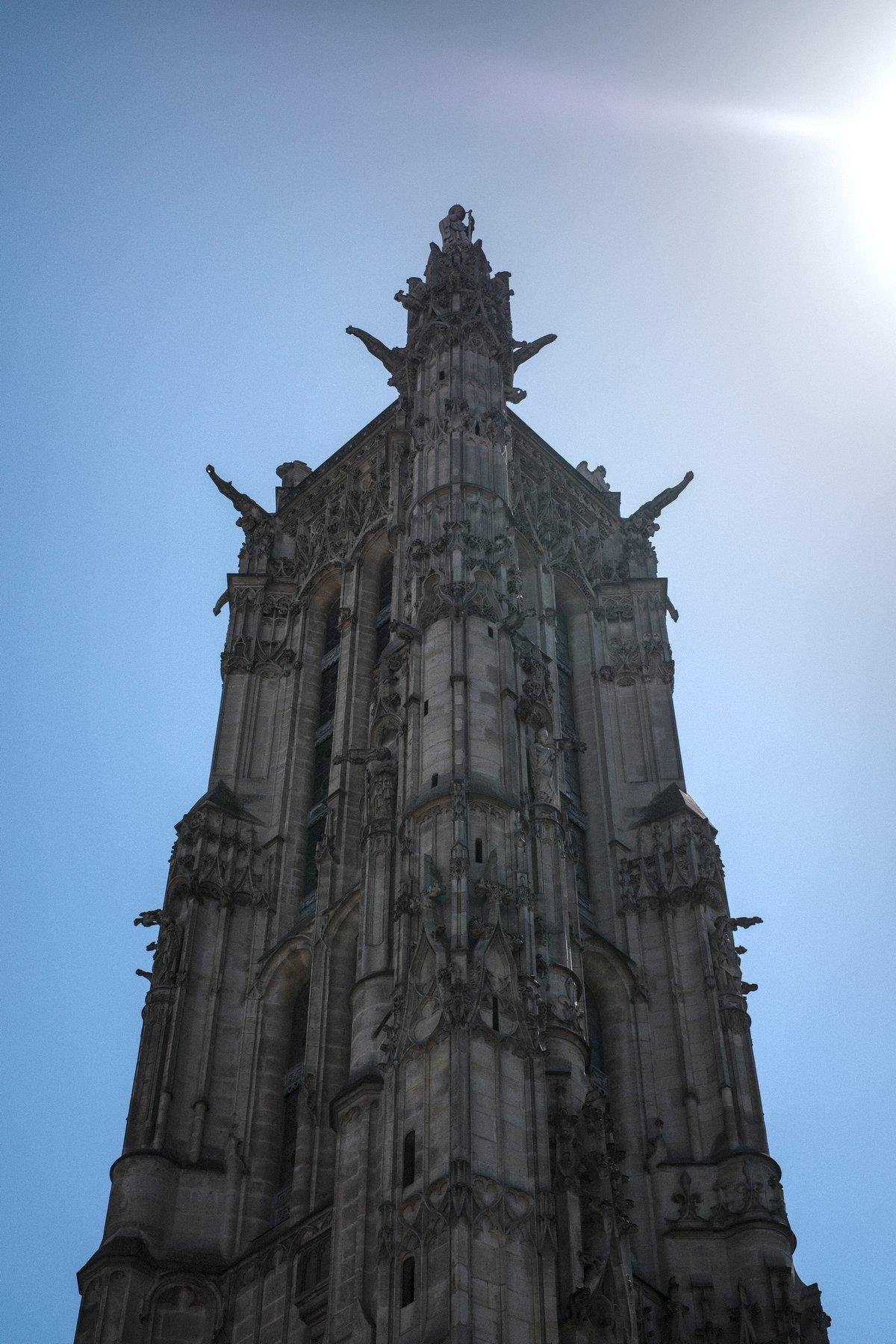 Photo verticale. Contre plongée sur la tour St Jacques à Paris, sous un ciel bleu qui se dégrade en blanc sur le coin supérieur droit, où le soleil brûle l’image