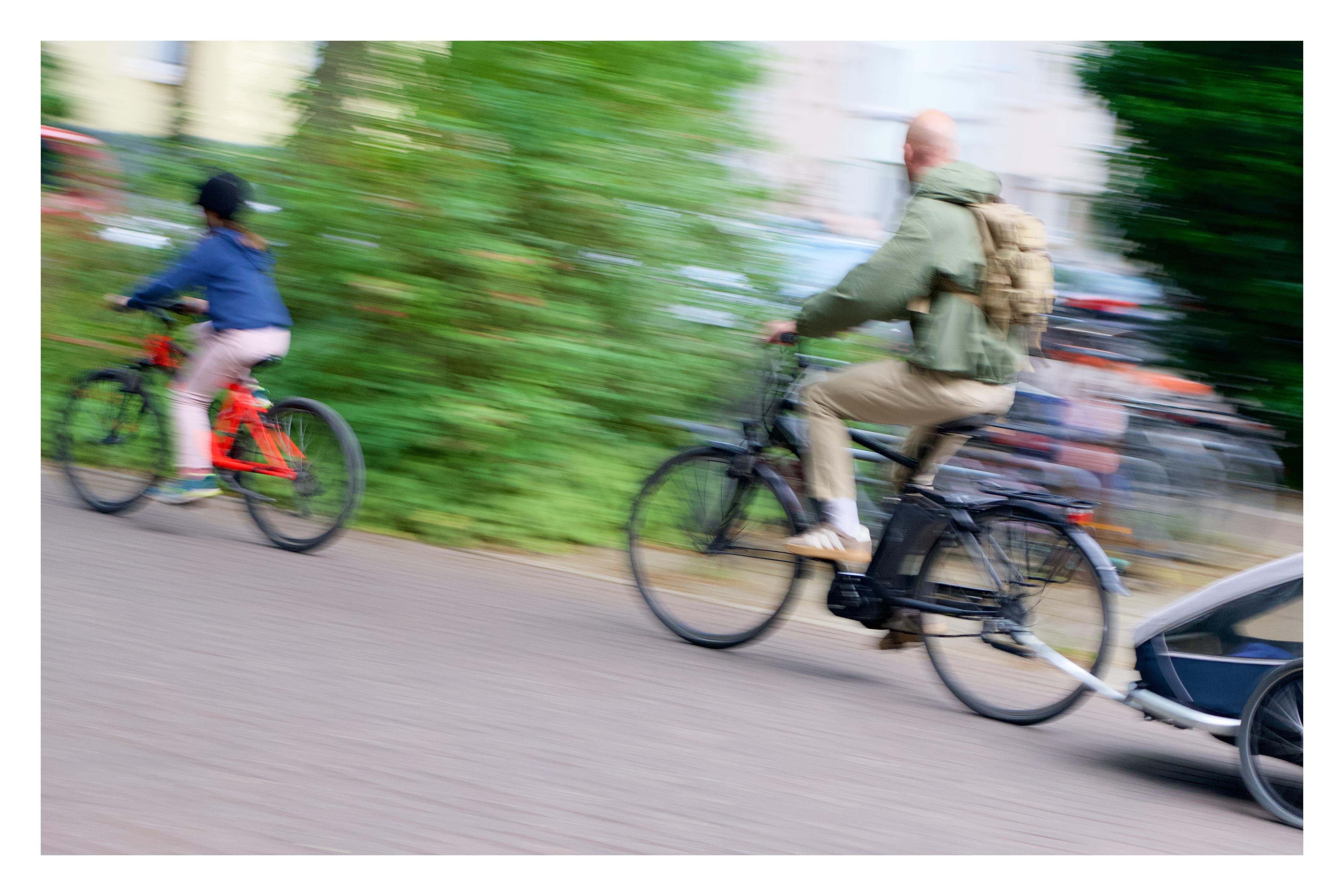 Ein Erwachsener mit Rucksack fährt auf einem Fahrrad mit Kinderanhänger hinter einem Kind auf rotem Fahrrad, alles in Bewegung mit verschwommenem Hintergrund.