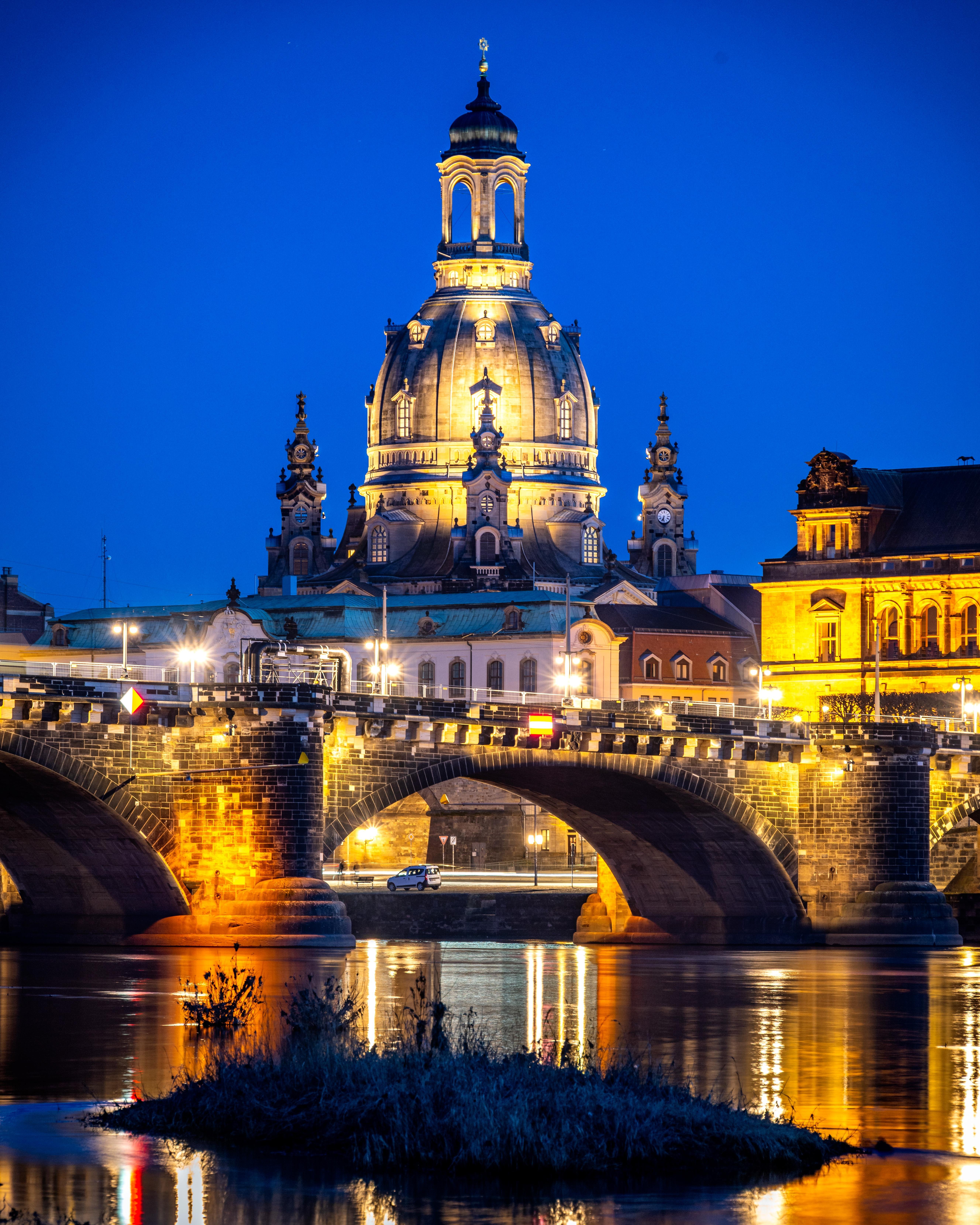 Illuminated domed cathedral and surrounding historic buildings at night behind a stone arch bridge, with streetlights along the bridge and golden reflections on the river in the foreground under a deep blue sky.