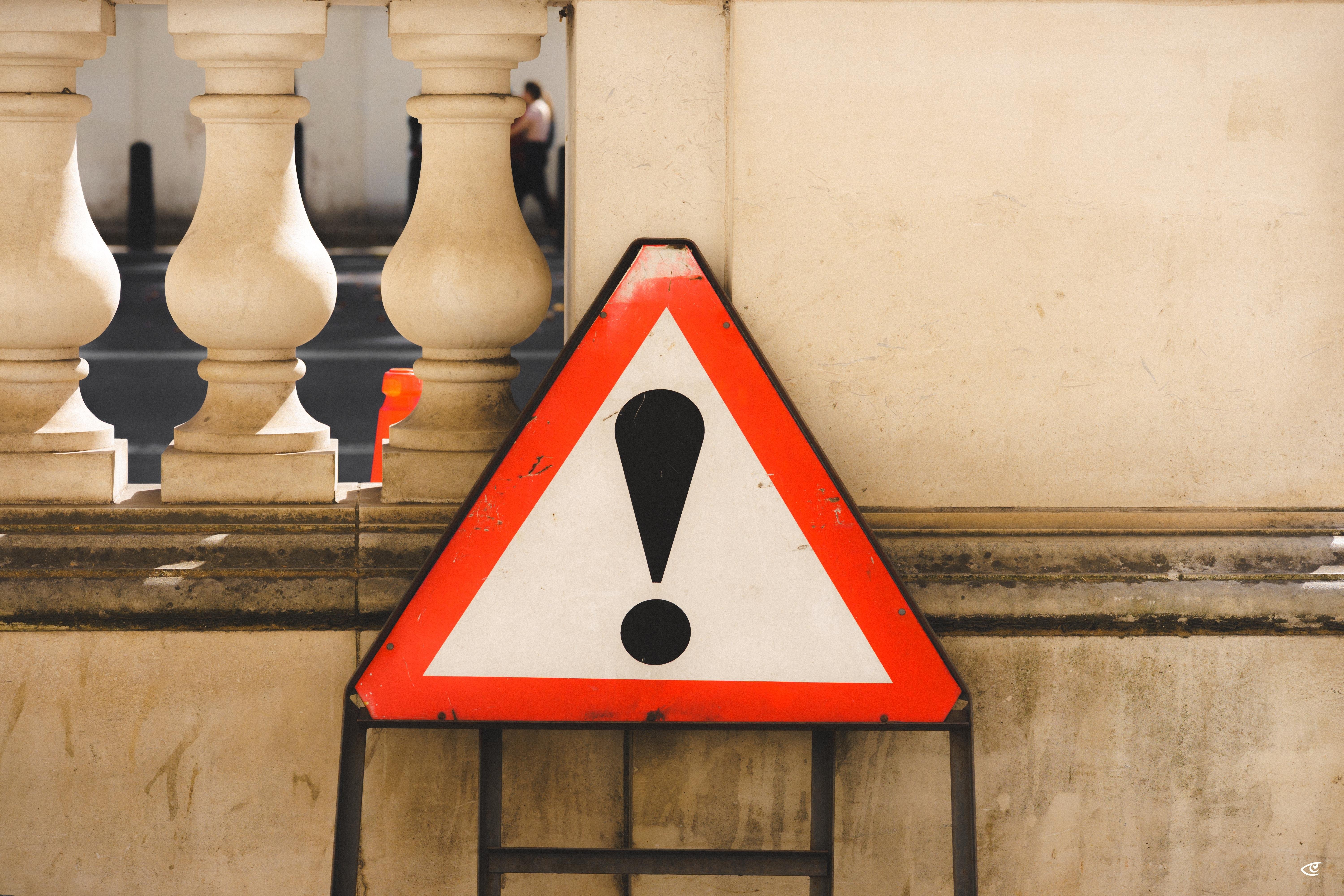 Red-and-white triangular warning sign with a black exclamation mark leaning against a beige stone wall beside a balustrade, with street and a blurred pedestrian in the background.