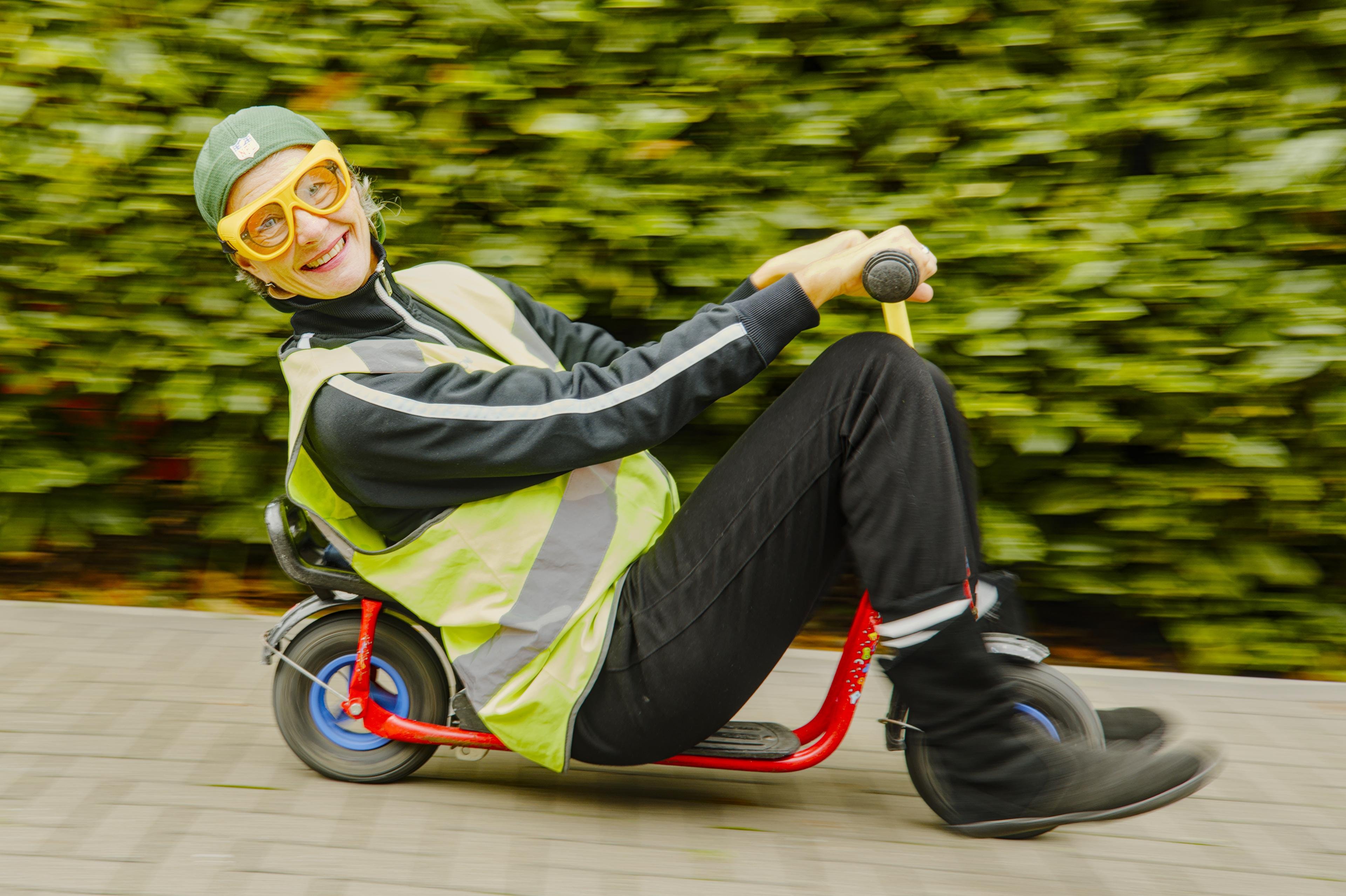 Person wearing a green cap and yellow goggles, riding a small red scooter with black wheels. Dressed in a reflective yellow vest and black pants, they are smiling while leaning back. Background features motion-blurred green foliage and a paved path, suggesting movement.
###
Eine Person mit grüner Mütze und gelber Schutzbrille fährt einen kleinen roten Roller mit schwarzen Rädern. Sie trägt eine reflektierende gelbe Weste und schwarze Hosen und lehnt sich lächelnd zurück. Im Hintergrund sind bewegungsunscharfe grüne Blätter und ein gepflasterter Weg zu sehen, die Bewegung suggerieren.