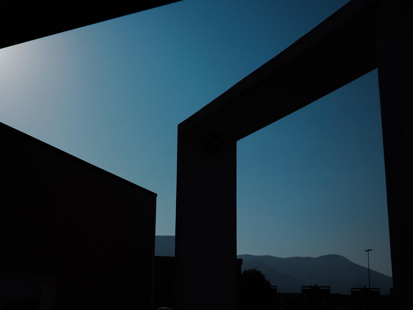 Montagne in lontananza sotto un cielo azzurro e limpido. In primo piano, elementi architettonici in silhouette disegnano forme geometriche.