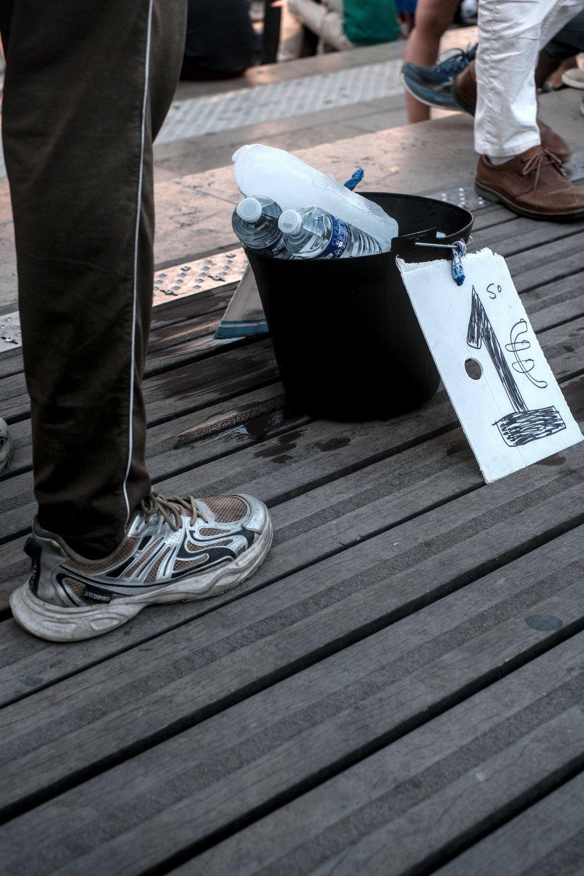 Photo verticale. Sur des planches en bois du début d’un pont piéton, un sceau avec un énorme pain de glace et des bouteilles d’eau minérales à vendre 1€ selon la pancarte posée devant. On voit des jambes de gens passants