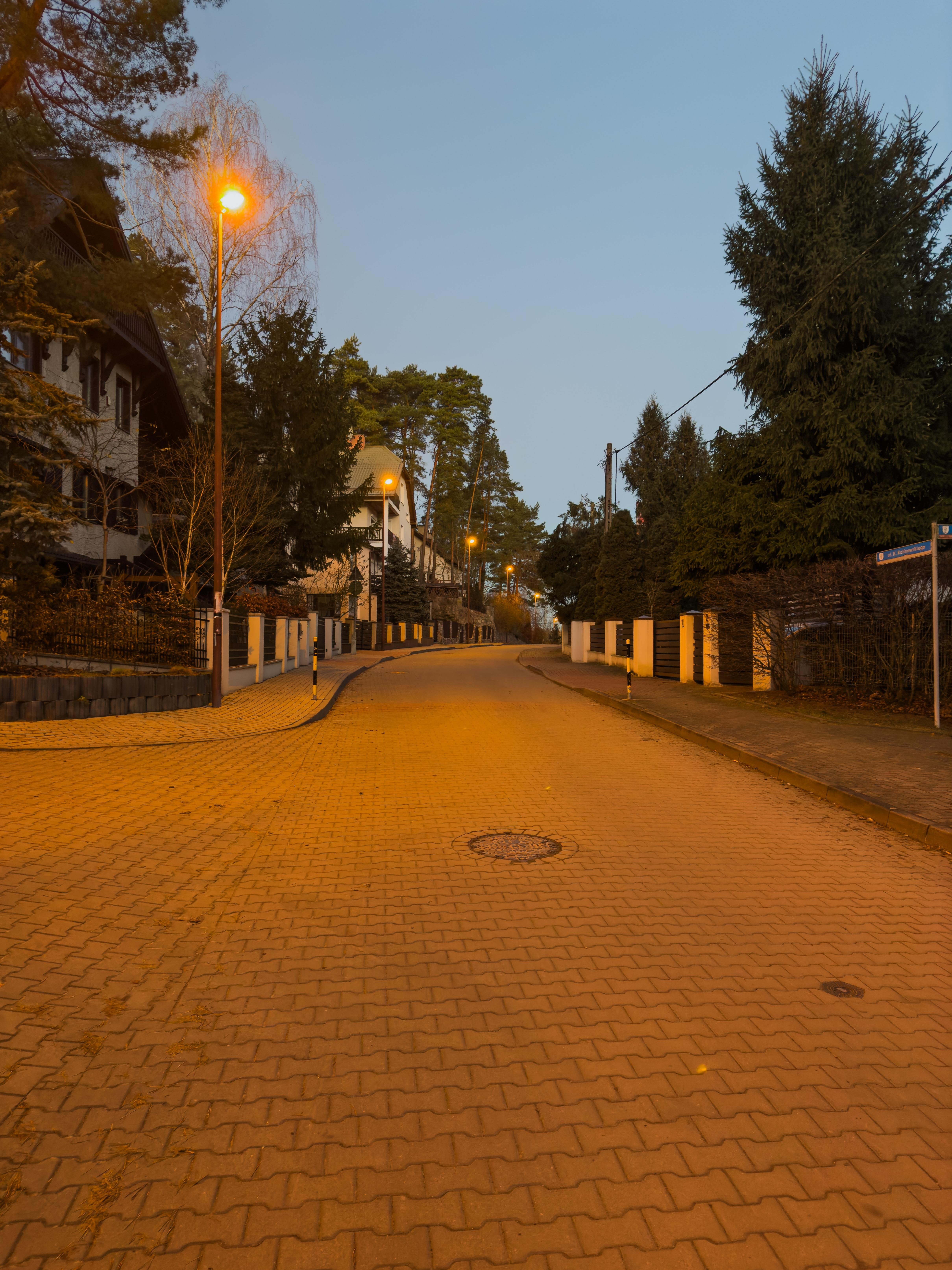 The image shows a quiet, residential street in the evening or early morning. The street is lined with trees on both sides and has houses or buildings visible. The streetlights are lit, casting a warm glow on the paved road. The area appears peaceful and suburban.