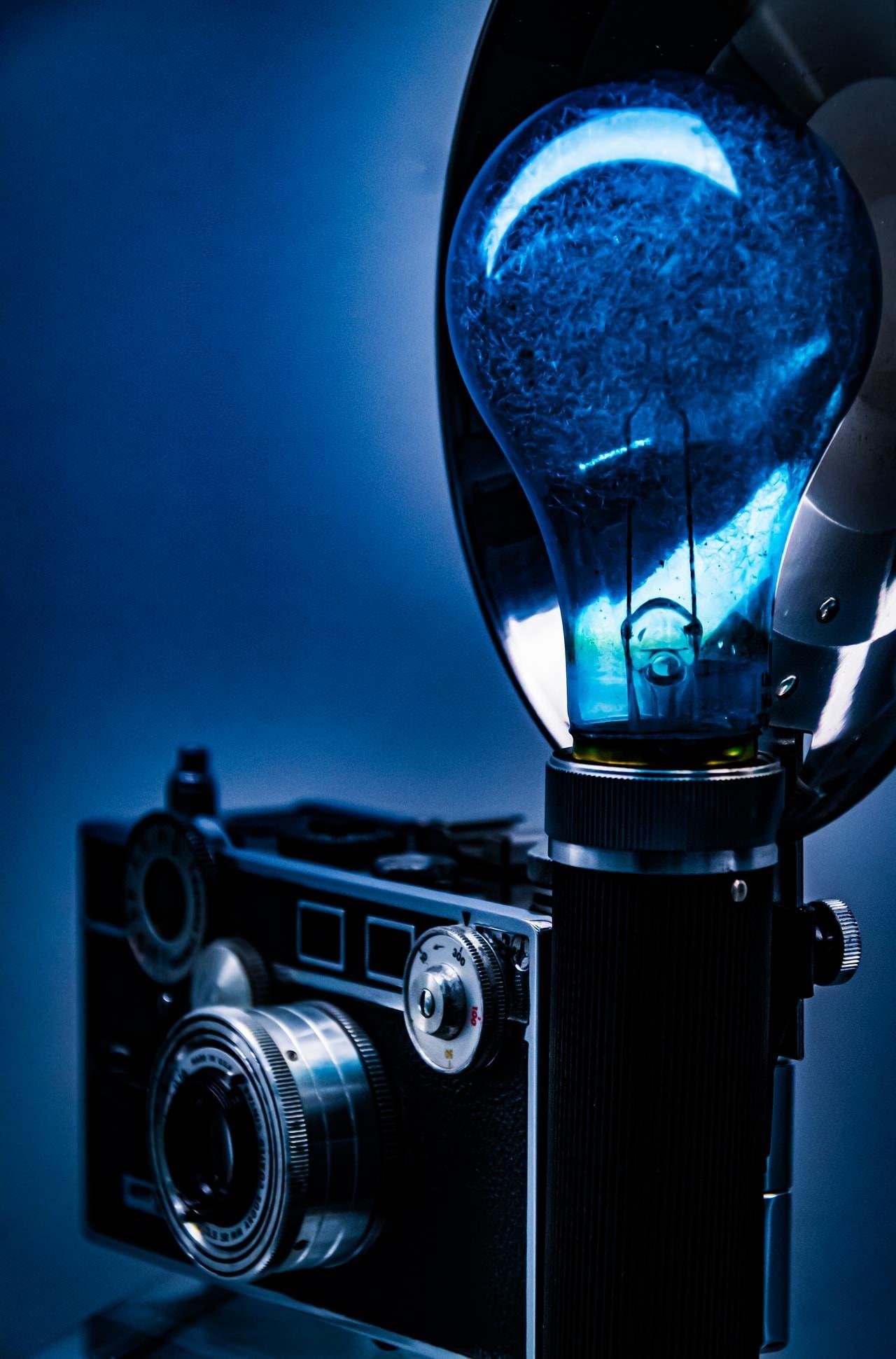 Close-up of a vintage film camera with a large blue-tinted flash bulb attached on the right, lit against a dark blue background with shallow depth of field.