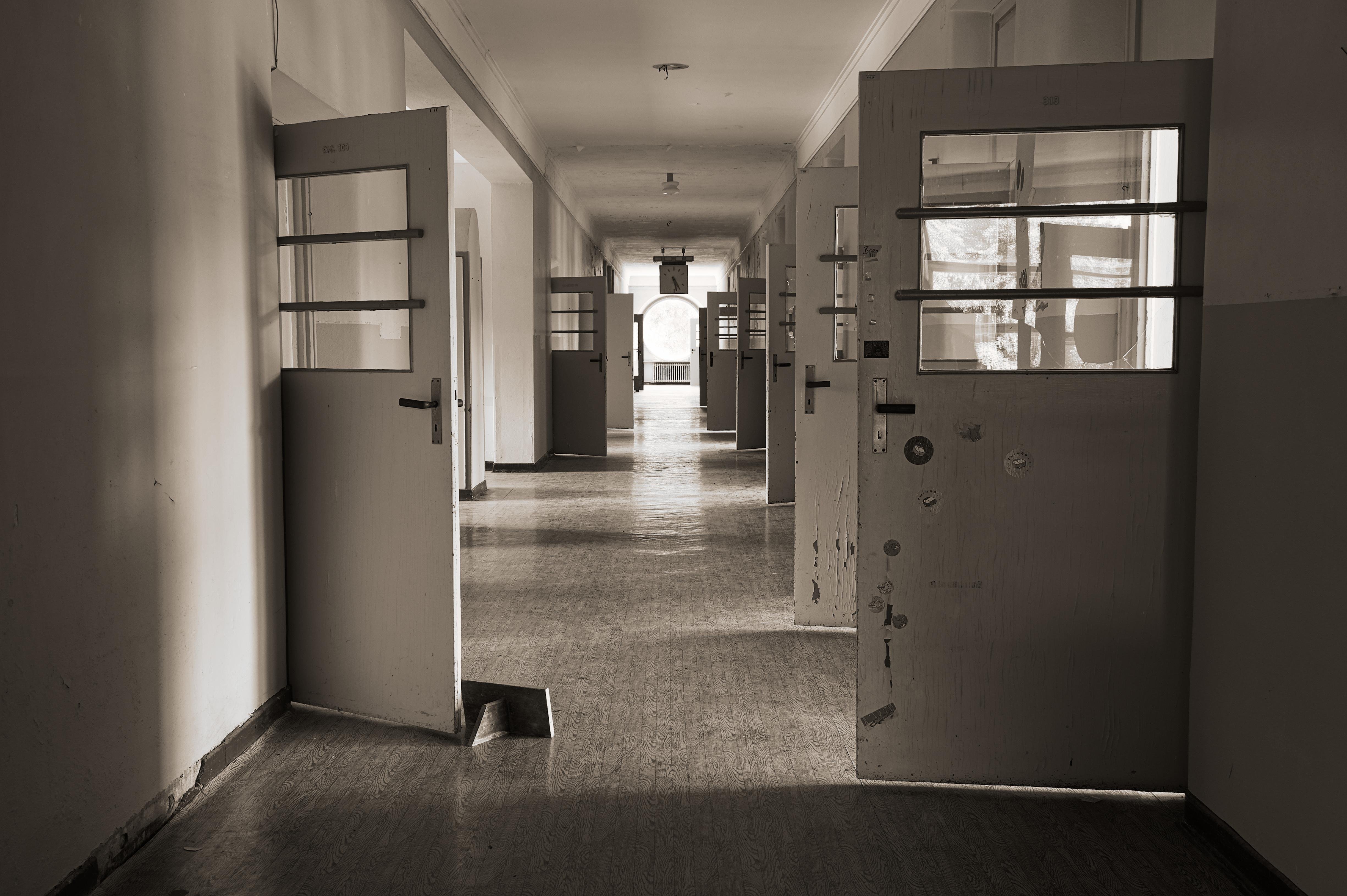 Dimly lit, monochromatic hallway lined with open doors on both sides. Each door has glass sections in the upper half, revealing bars. The floor is worn, and the corridor leads to a distant bright window, casting light along the path. Ceiling lights are evenly spaced, adding to the sparse illumination.
###
Schwach beleuchteter, einfarbiger Flur mit offenen Türen auf beiden Seiten. Jede Tür hat in der oberen Hälfte Glasabschnitte, die Gitterstäbe freilegen. Der Fußboden ist abgenutzt, und der Gang führt zu einem fernen, hellen Fenster, das Licht auf den Weg wirft. Die Deckenleuchten sind gleichmäßig verteilt, was die spärliche Beleuchtung noch verstärkt.
