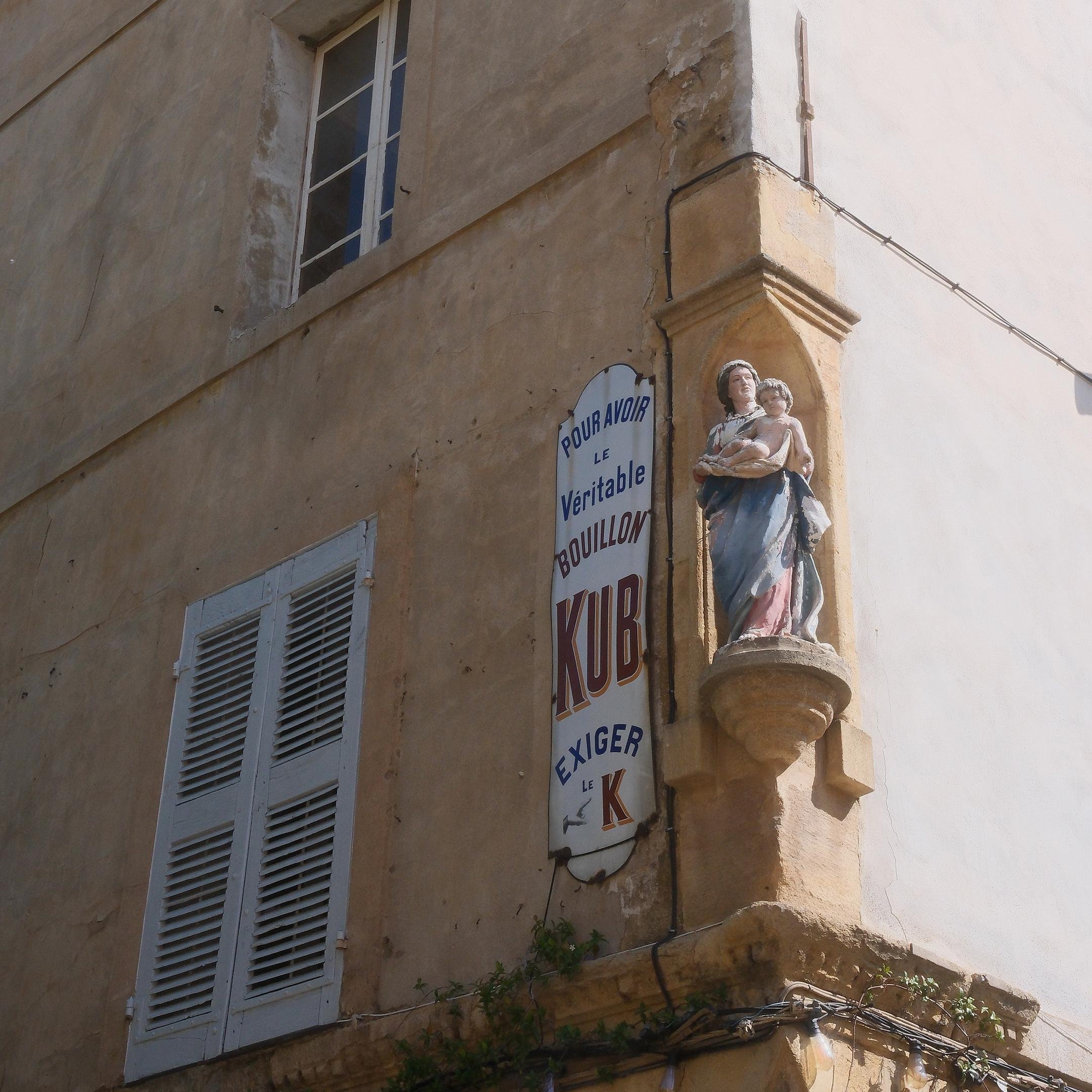 Photo carrée. Un coin de rue avec dans l’angle d’un immeuble, une statue colorée d’une femme en robe bleue portant un enfant nu dans ses bras, et à gauche, une vieille plaque de réclame pour les bouillon KUB