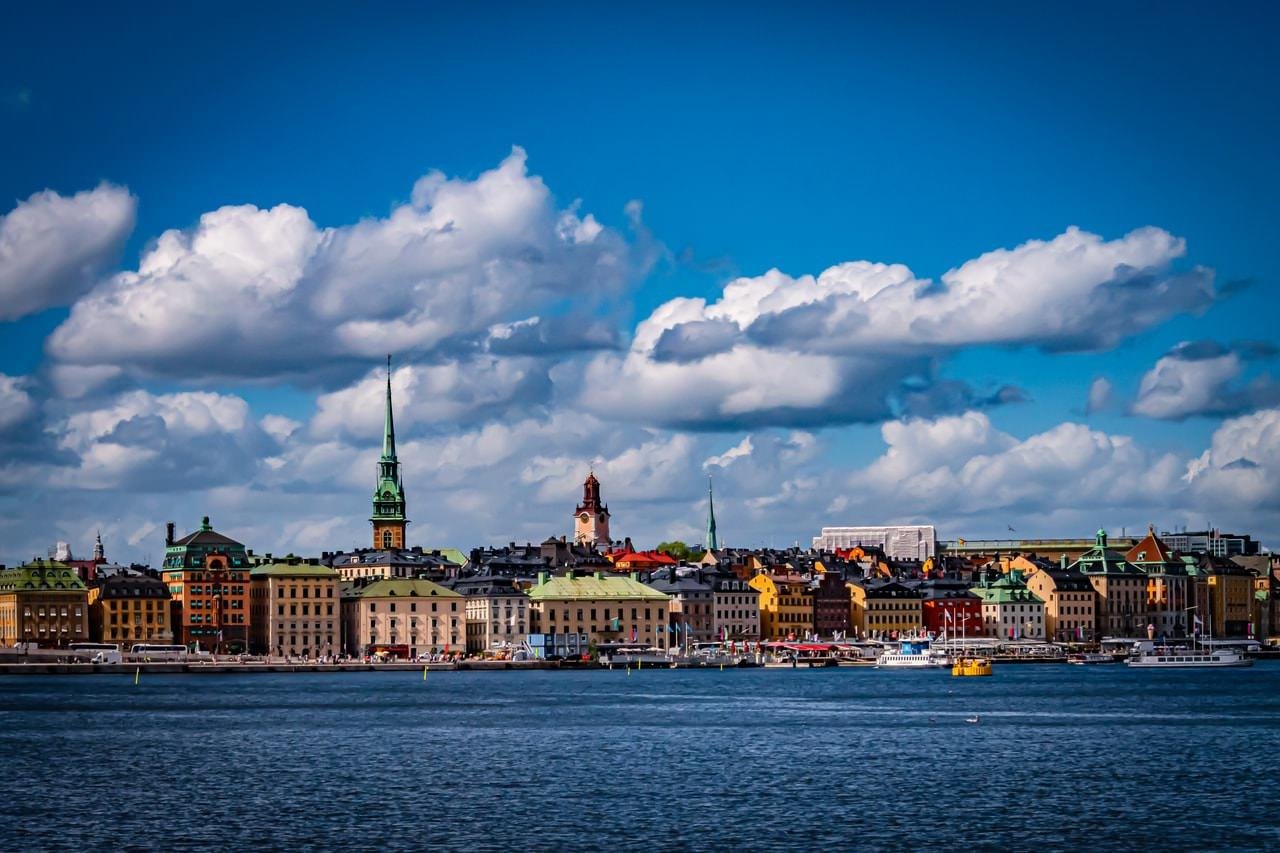 A cityscape of Stockholm with colorful historic buildings along a waterfront under a bright blue sky. Several church steeples rise prominently, surrounded by cumulus clouds. Boats are visible in the water, and the overall scene appears vibrant and lively.