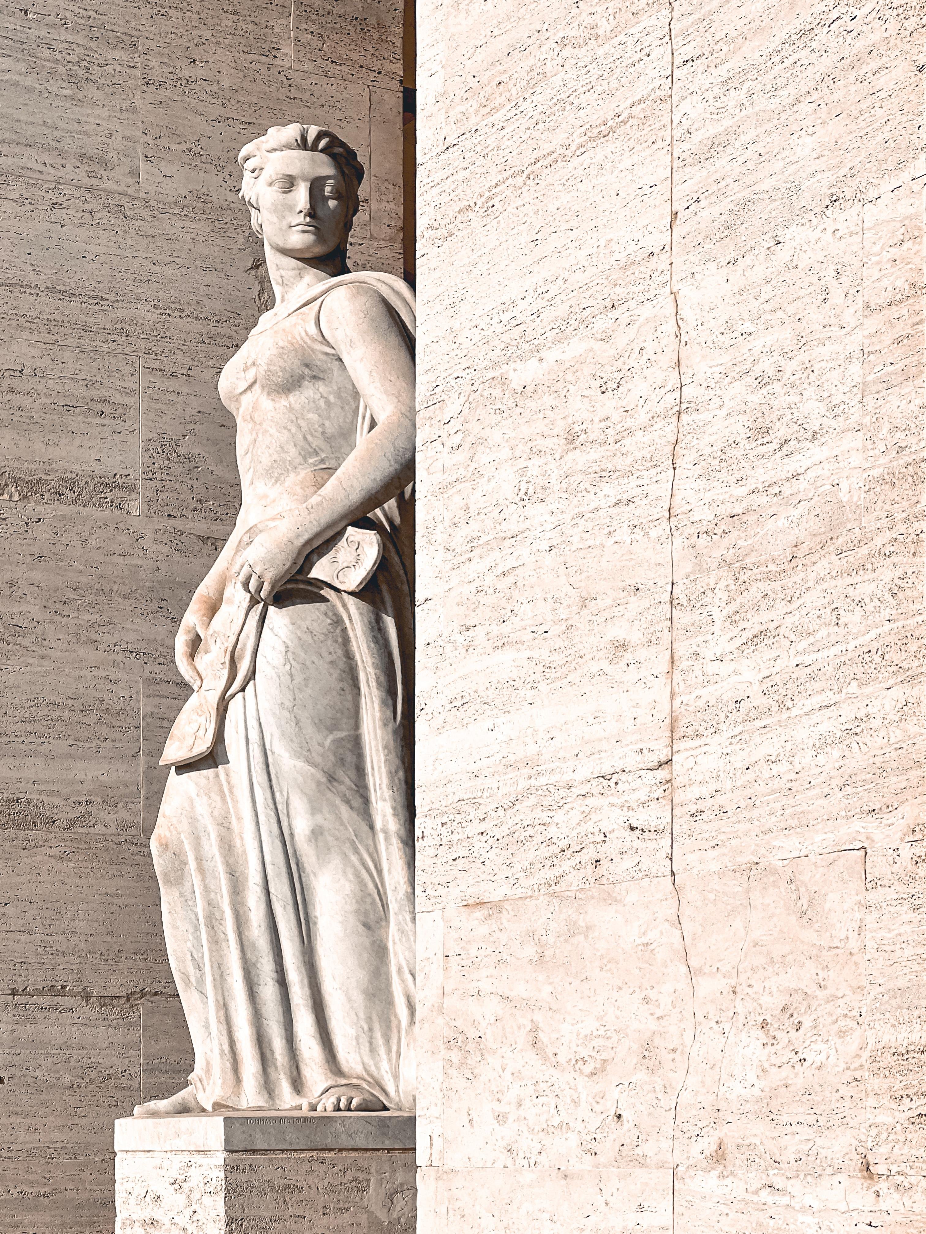 Statua in marmo di una figura classica, probabilmente rappresentante una persona dell’antichità o della mitologia. La statua è posta contro una parete di marmo, evidenziando i dettagli scolpiti e le pieghe del drappeggio.