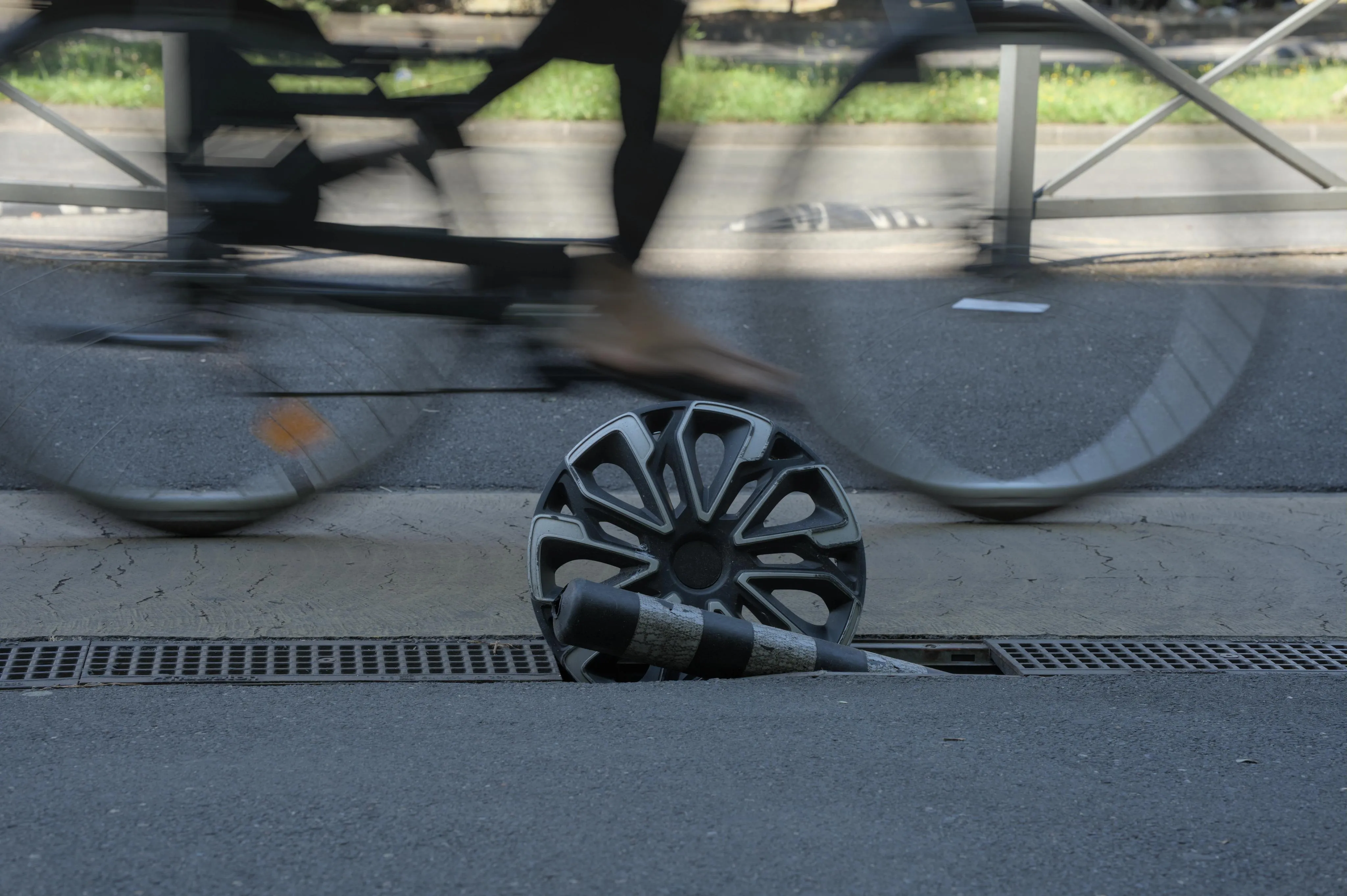 Photograph of the rim of an automobile wheel planted in the middle of the drain in a sidewalk, along with a broken cylindrical roadblock plot. The wheel rim has traces of wear and tear. The sidewalk is in the shade. Behind the lost rim in the drain, part of a bicycle (up to its wheels) is visible in motion blur.