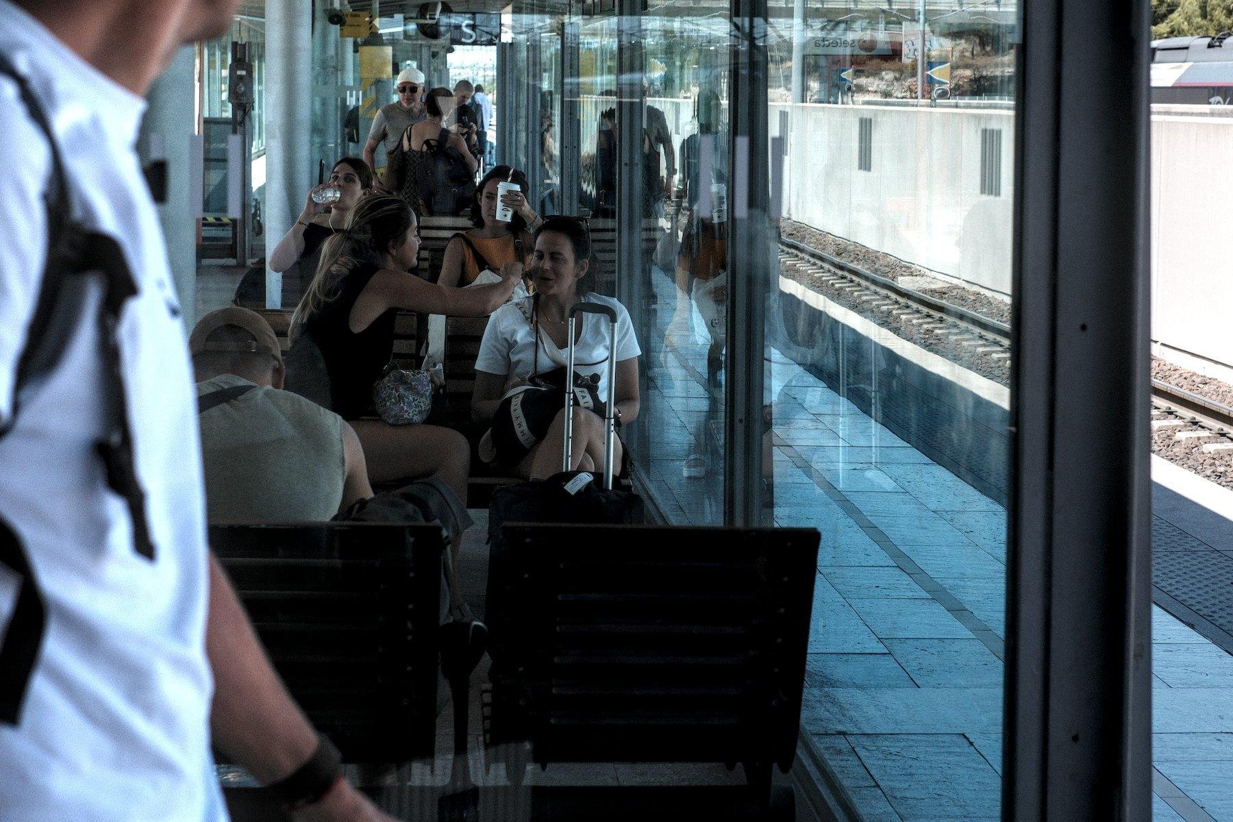 Photo horizontale. Dans une zone d’attente encadrée par de vitres sur un quai de gare, 2 femmes assise côte à côte. Celle de gauche repoudre le visage de son amie à droite
