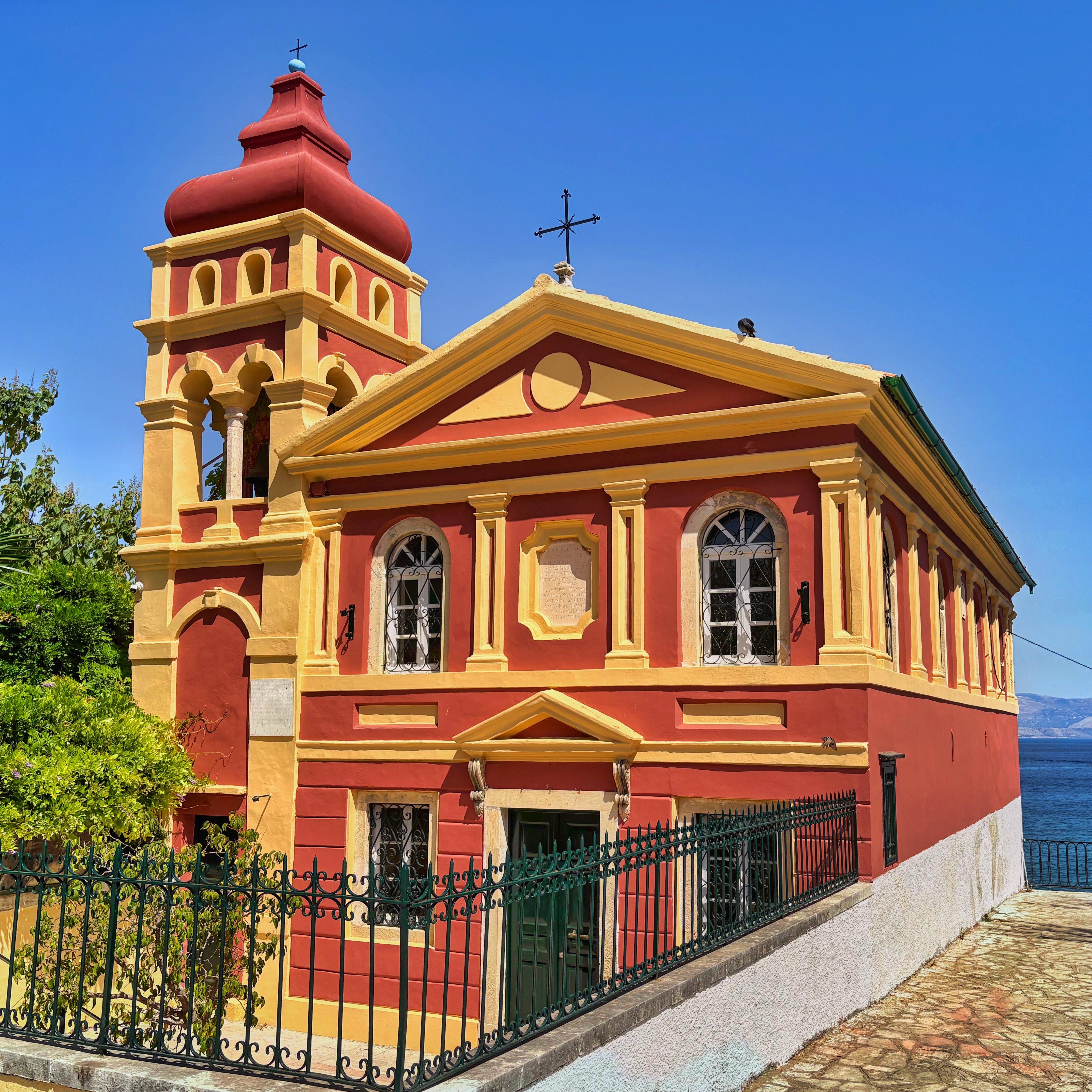 Vibrantly colored church with a red and yellow facade, featuring arched windows and a bell tower topped with a cross. The building is surrounded by a black metal fence, with green foliage visible in the background. Clear blue sky and distant water provide a scenic backdrop.