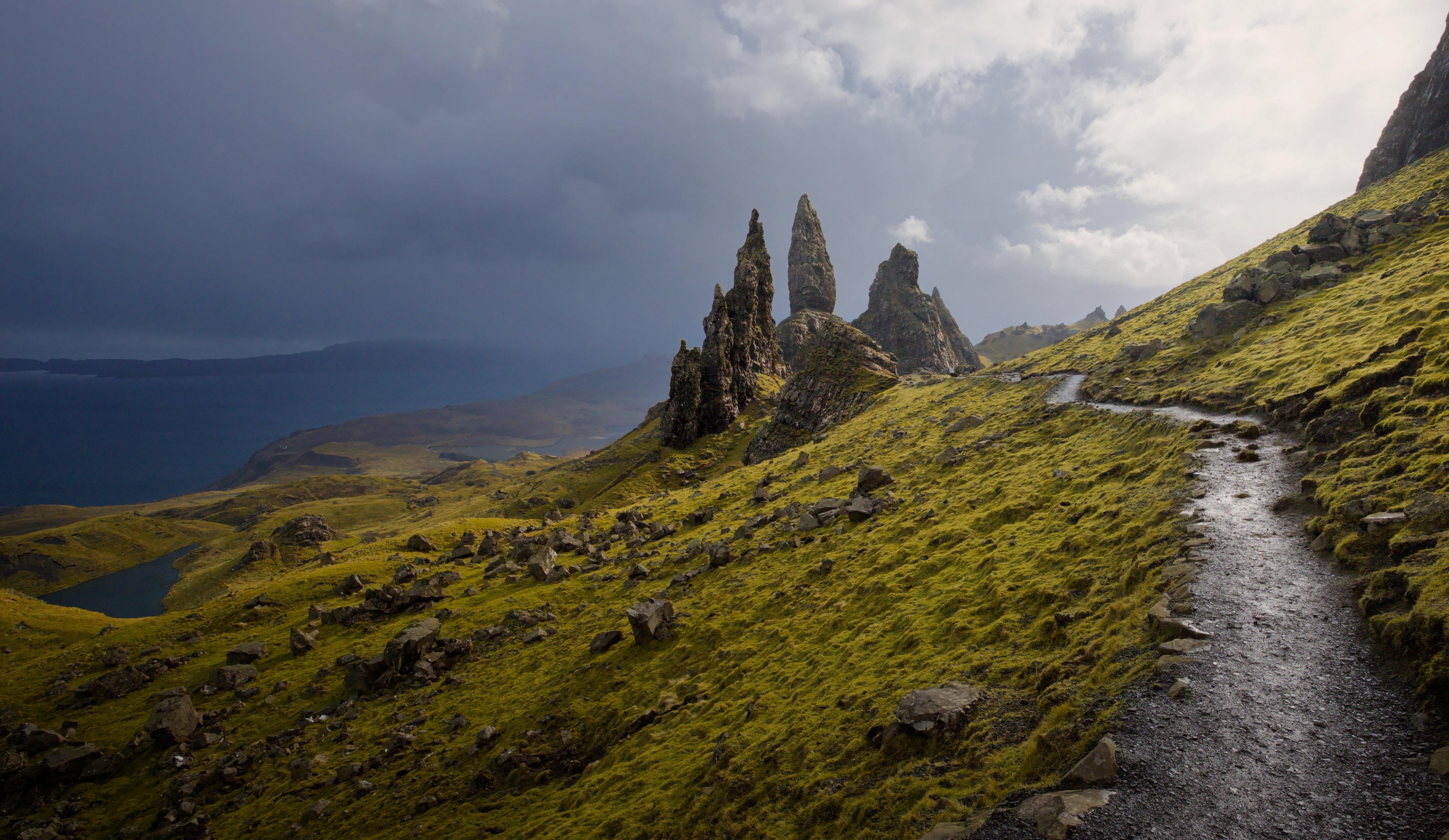 Narrow gravel path along a steep grassy hillside leading toward jagged rock spires on a ridge. Dark clouds loom overhead.