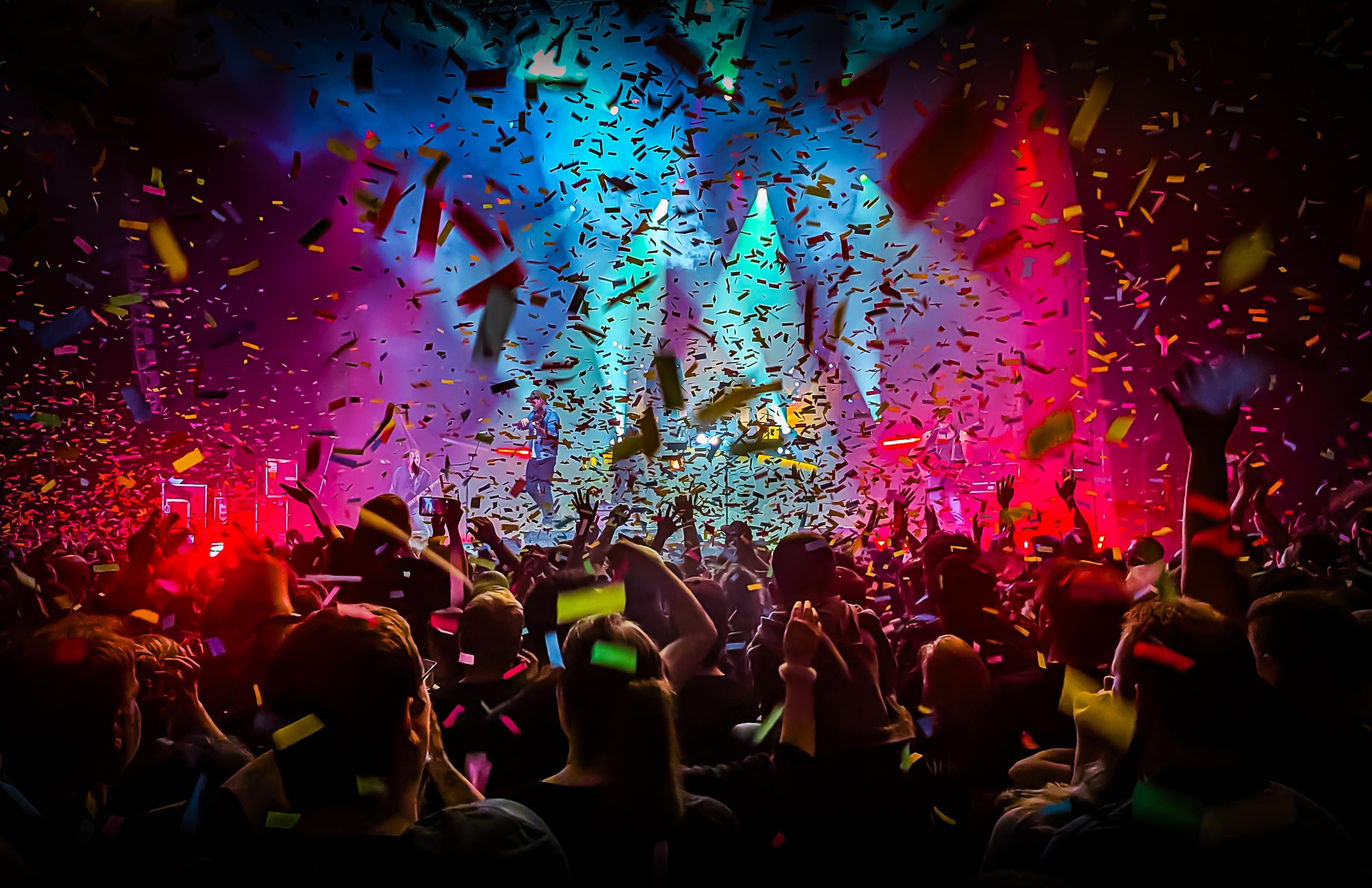 Crowd at a concert with raised hands facing a brightly lit stage, blue and pink spotlights, and multicolored confetti falling across the scene.