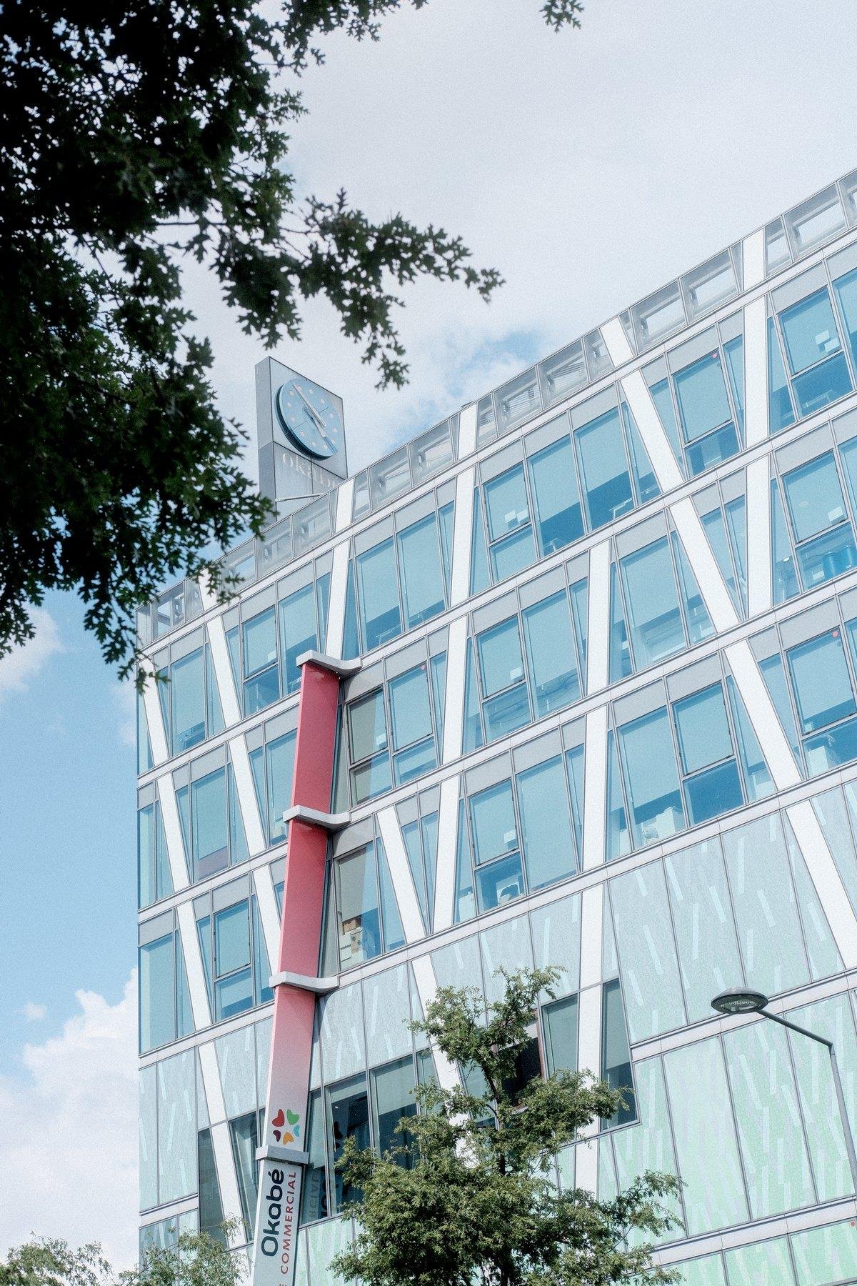 Photo verticale. Une horloge analogique au sommet d’un bâtiment moderne fait de verre et d’acier, cadrée par le feuilage d’un arbre