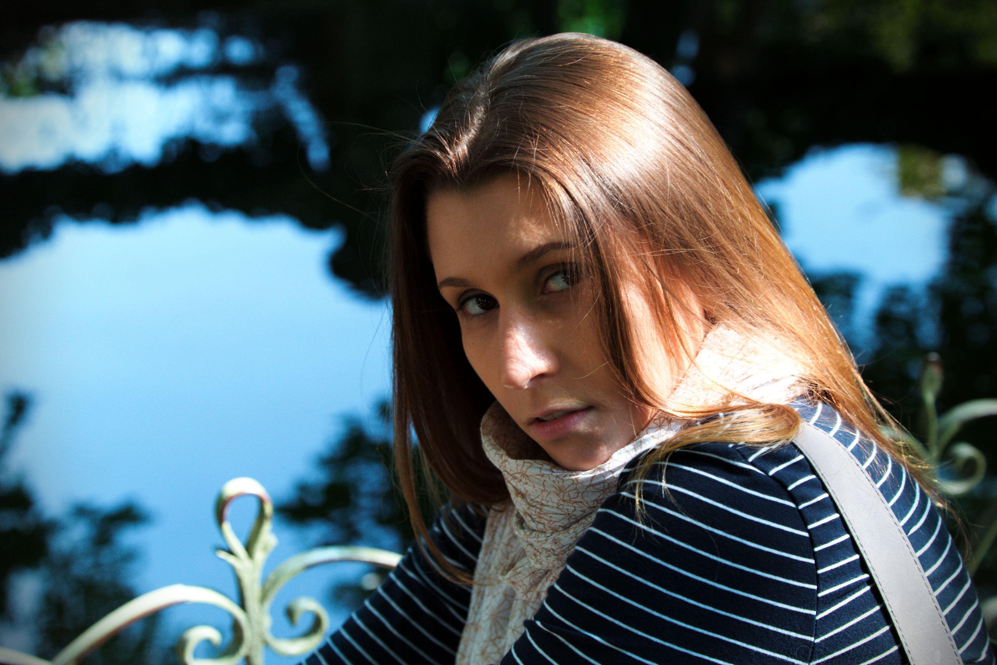 Photo format paysage, montrant le portrait d’une jeune femme brune au cheveux long, portant un haut bleu rayé de blanc, un foulard blanc à motif fleuris marron/orangé, tournant légèrement la tête vers la caméra et les yeux en coin vers l’objectif, avec un regard intense