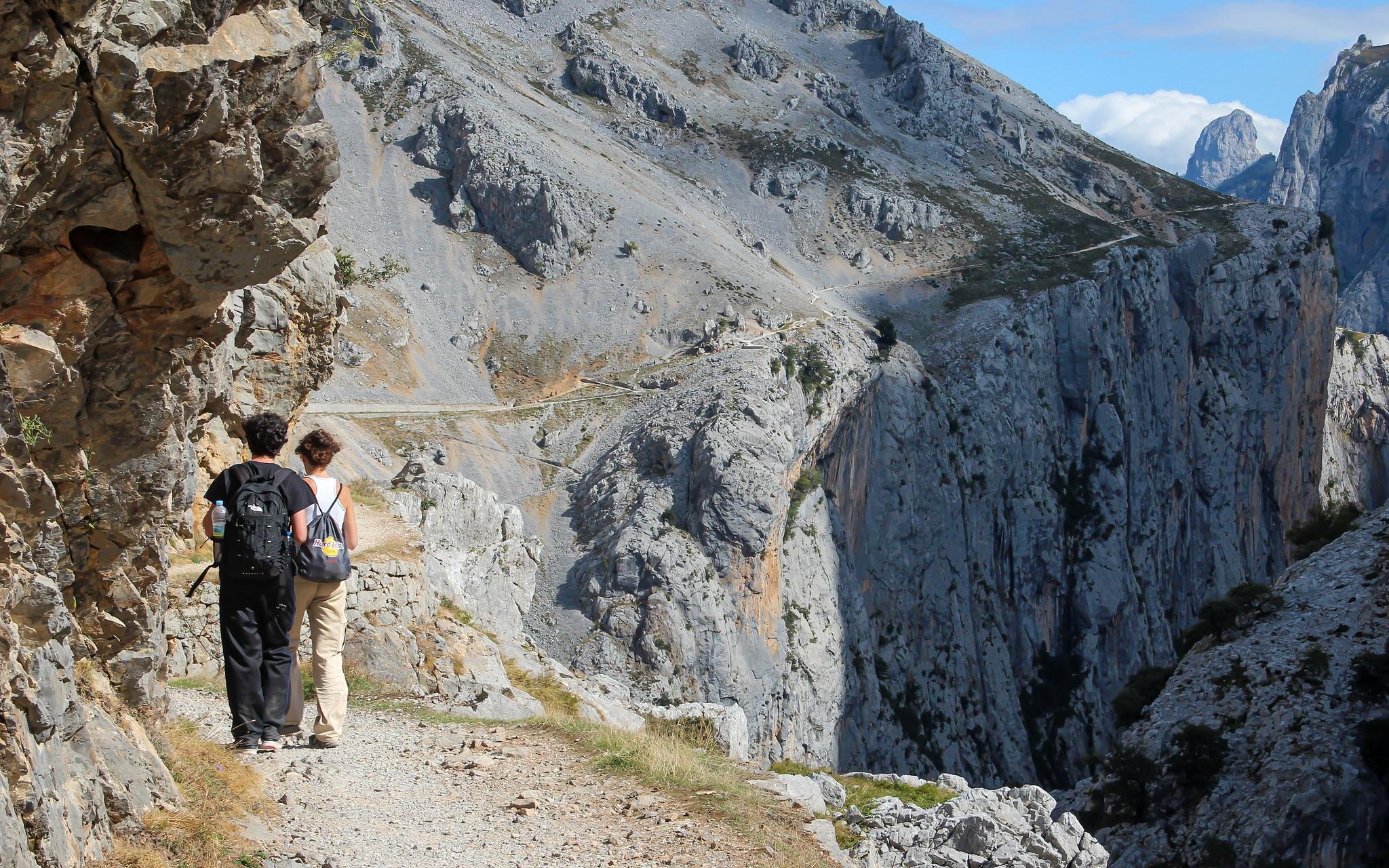 The image shows two people hiking on a narrow trail through rocky, mountainous terrain. They are walking along a path that cuts through the rugged landscape, with steep cliffs and rocky outcroppings surrounding them. The setting appears to be a scenic and challenging hiking area.