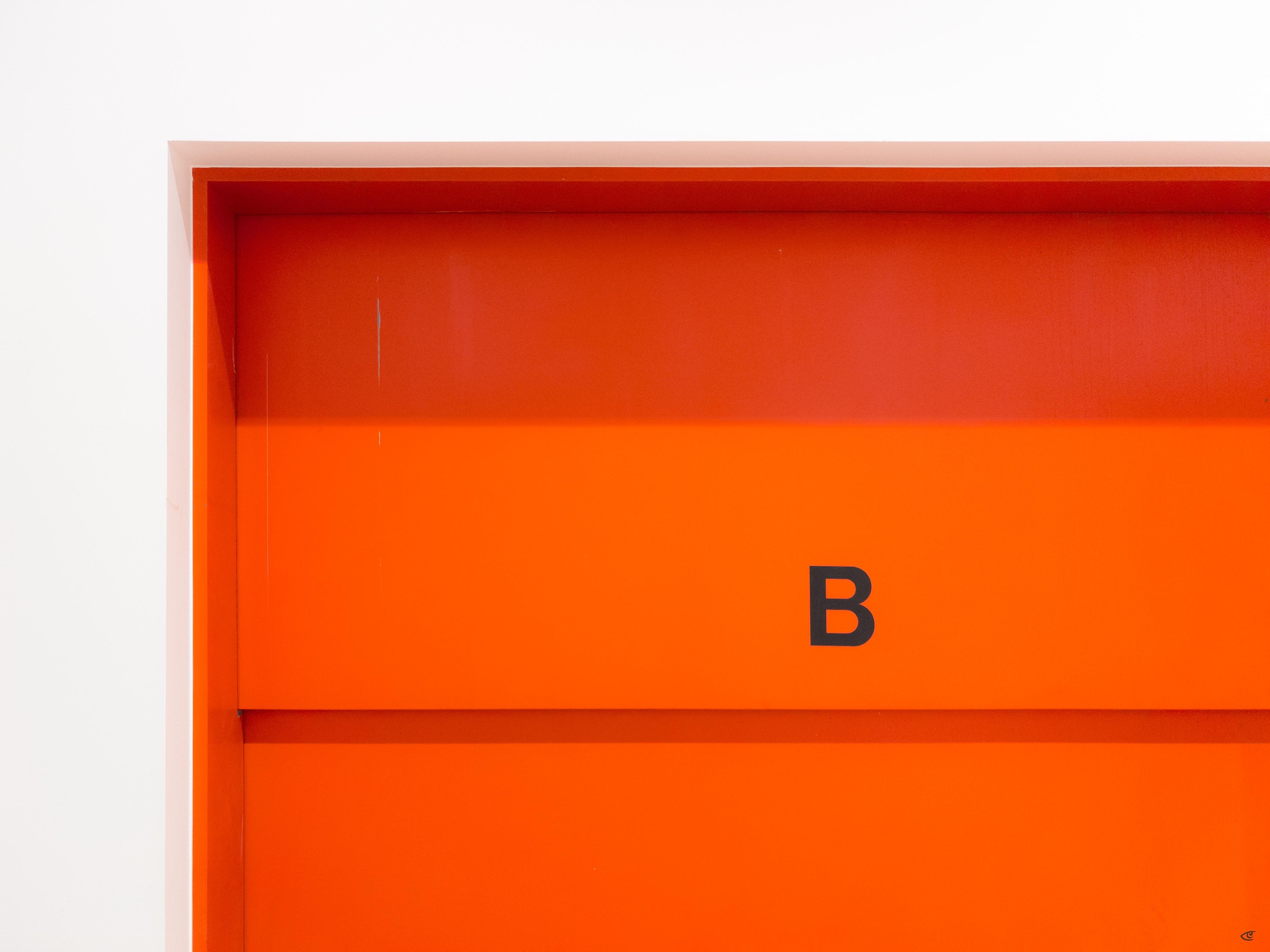 Orange wall or door panel with a recessed rectangular border and the black letter “B” centered slightly right, set against a white edge on the left.