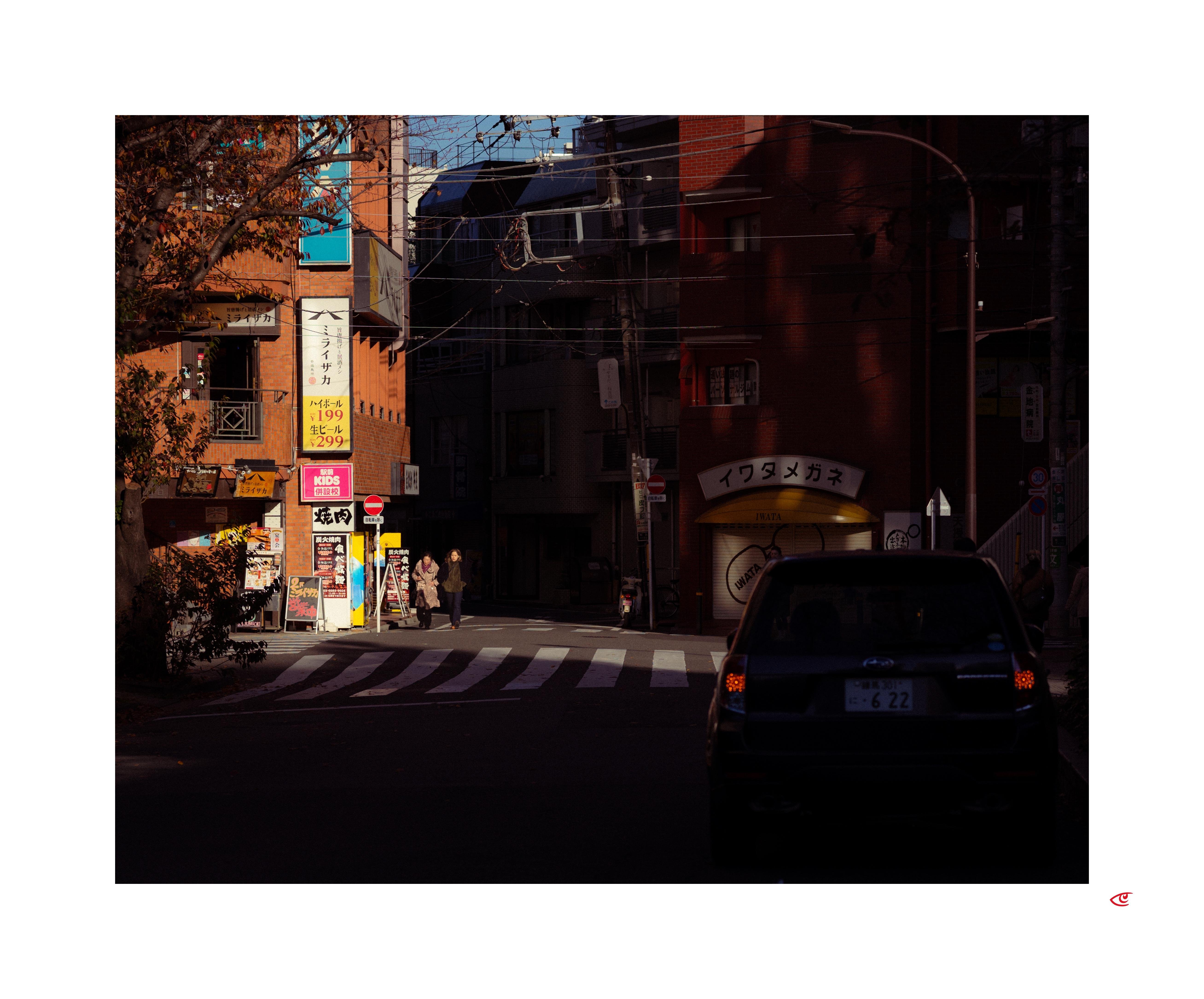 Urban street intersection with a zebra crosswalk, a dark car in the foreground at right, and a few pedestrians near storefronts. Sunlit building facades with Japanese signs on the left contrast with shadowed buildings in the center and right; overhead wires and a tree frame the scene.