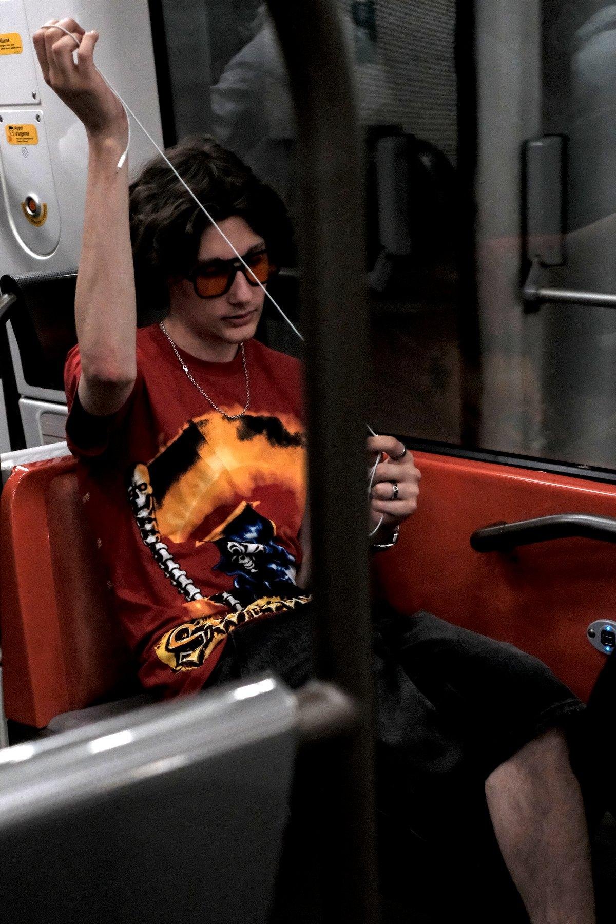 Photo verticale. Dans une rame de métro, un jeune homme assis autour des parois oranges de la ligne 14, t-shirt coloré rouge avec une illustration à dominante orange et noir, petite chaînette autour du cou, lunettes noire aux verre coloré en rouge, entrain de finir de déméler le câble blanc de ses écouteurs en tirant une extrémité vers le haut avec sa main droite