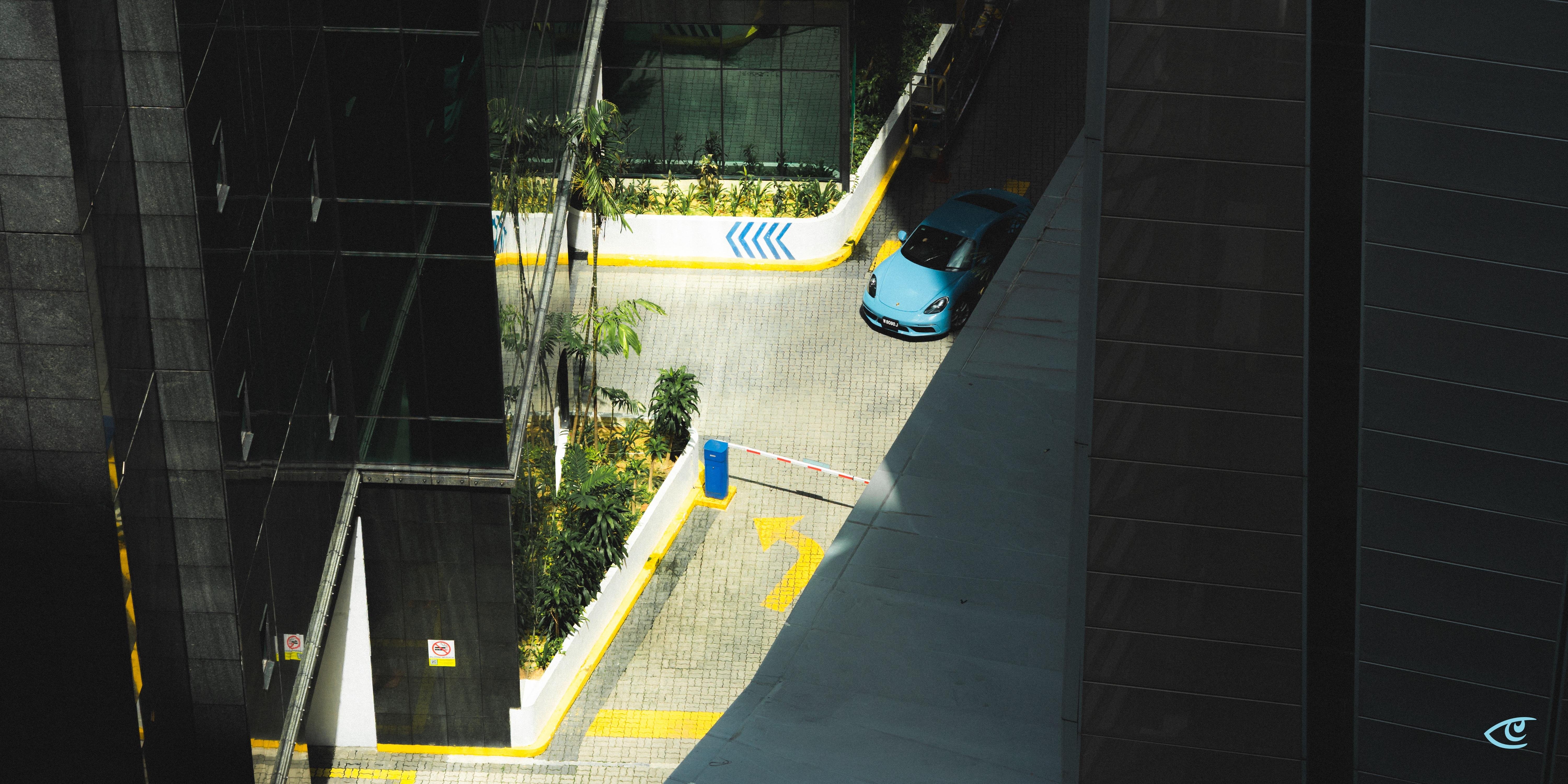 Overhead view of a paved driveway between tall dark buildings, with a blue car near the top right, a red-and-white barrier gate across the lane, and yellow painted arrows and curbs alongside small landscaped plants.