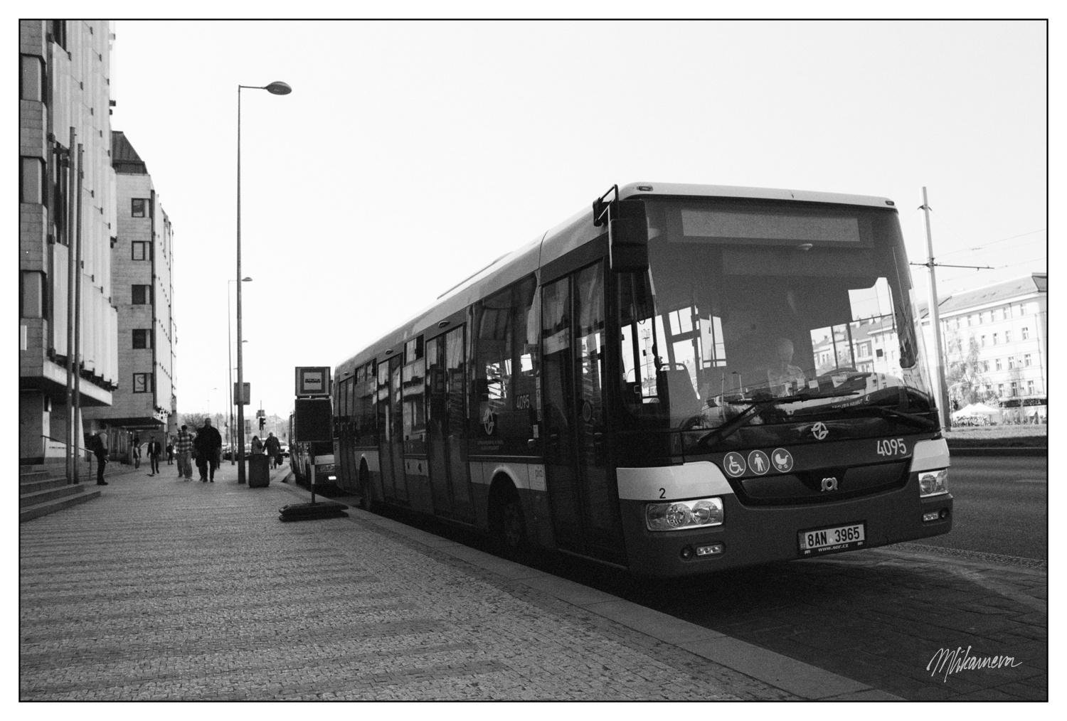 Black and white image of a city street with a large bus parked at a bus stop. The bus displays the number 4095 and has a visible license plate, 8AN 3965. Several pedestrians walk along the sidewalk bordered by tall buildings. Streetlights and tram lines are visible in the background.