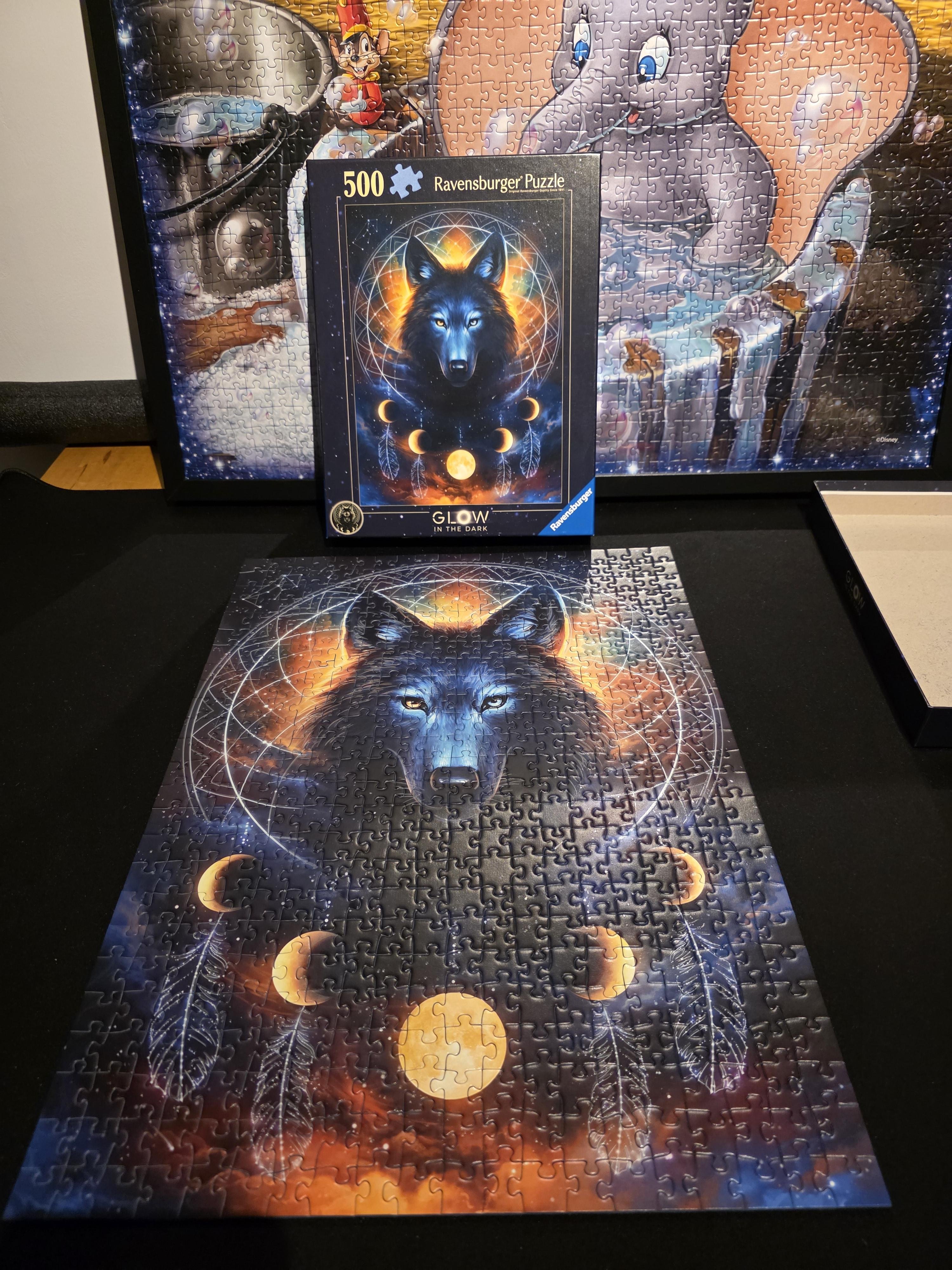 The image shows the fully completed Ravensburger Wolfmoon glow-in-the-dark puzzle laid neatly on a black surface. The 500-piece puzzle depicts a majestic blue-and-black wolf emerging from swirling cosmic light, its piercing icy eyes glowing against an ethereal backdrop of orange, gold, and deep midnight tones. Surrounding the wolf are delicate geometric patterns, feathers, and a sequence of glowing moon phases, giving the artwork a mystical, celestial feel. Behind the finished puzzle stands the matching Ravensburger puzzle box, showing the same artwork. In the background, another framed puzzle can be seen—an illustration featuring Disney’s Dumbo—its pastel colors and whimsical style contrasting with the dark, magical atmosphere of the Wolfmoon scene. The overall setting highlights the satisfaction of a freshly completed project and its vibrant, otherworldly design.