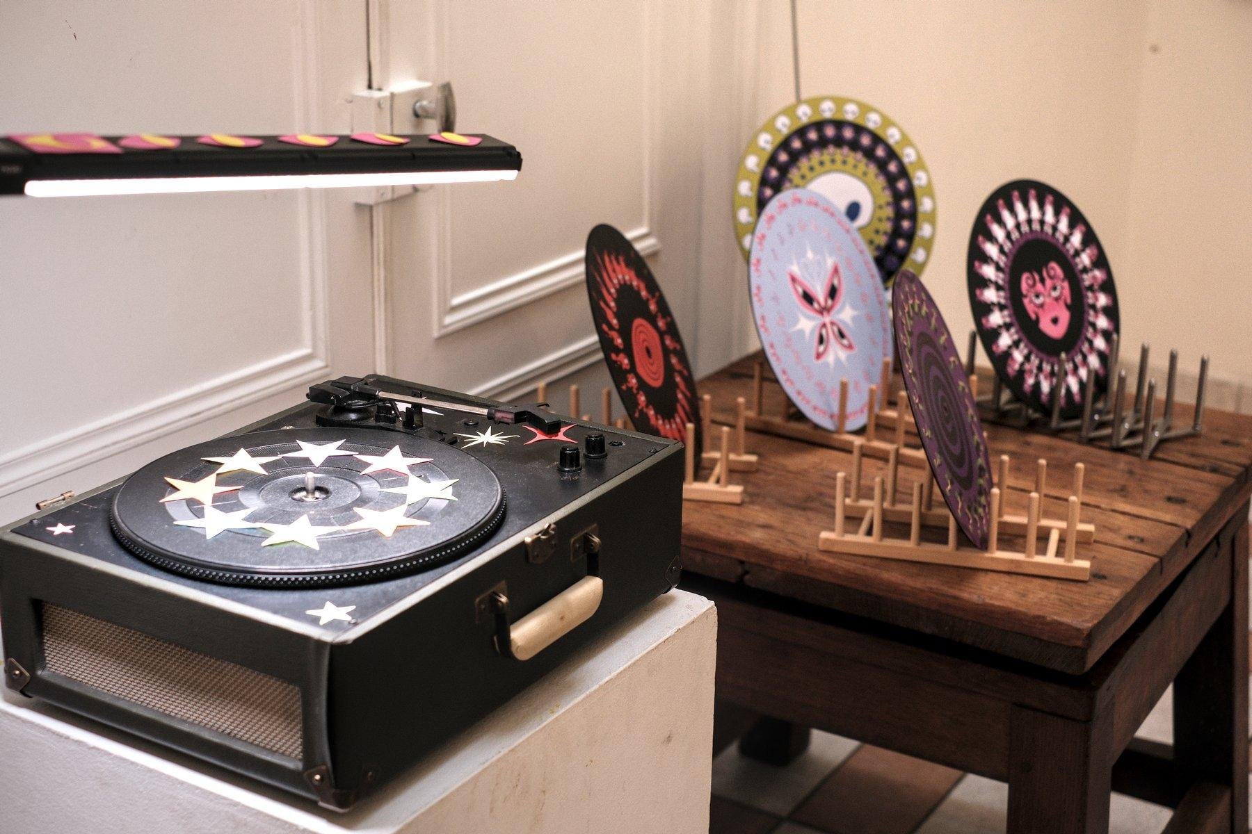 Photo horizontale. Au premier plan, une platine vinyle décorée d’étoiles en papier collées dessus et éclairée par une forte lampe au dessus, au second plan, 5 vinyls dont la face avant est totalement décorée d’œuvres différentes, posés sur des stands.