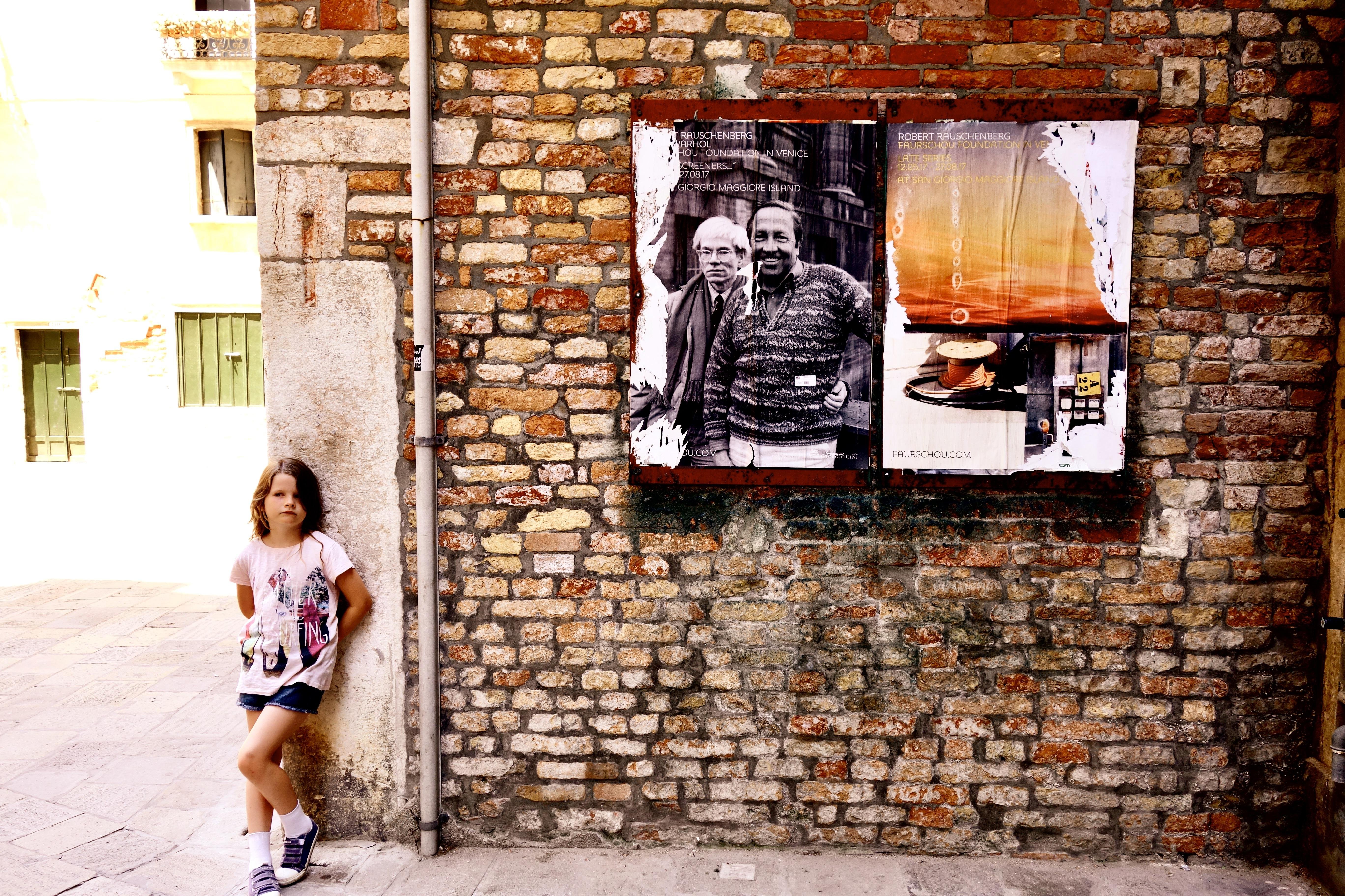 Young girl in a pink T-shirt and denim shorts leaning against a stone corner on the left, with a large brick wall behind her displaying two posters—one black-and-white portrait of two men and one orange-toned image—above a cobblestone walkway.