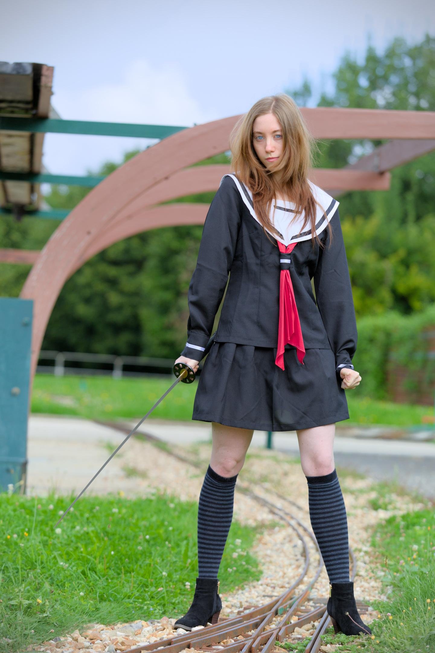 Une rousse habillée en écolière japonaise bleu marine avec un nœud rouge, debout, tenant un katana de la main droite, poing gauche serré, les jambes écartées bien plantées au sol, regardant vers la caméra avec ses yeux d’un bleu clair remarquable