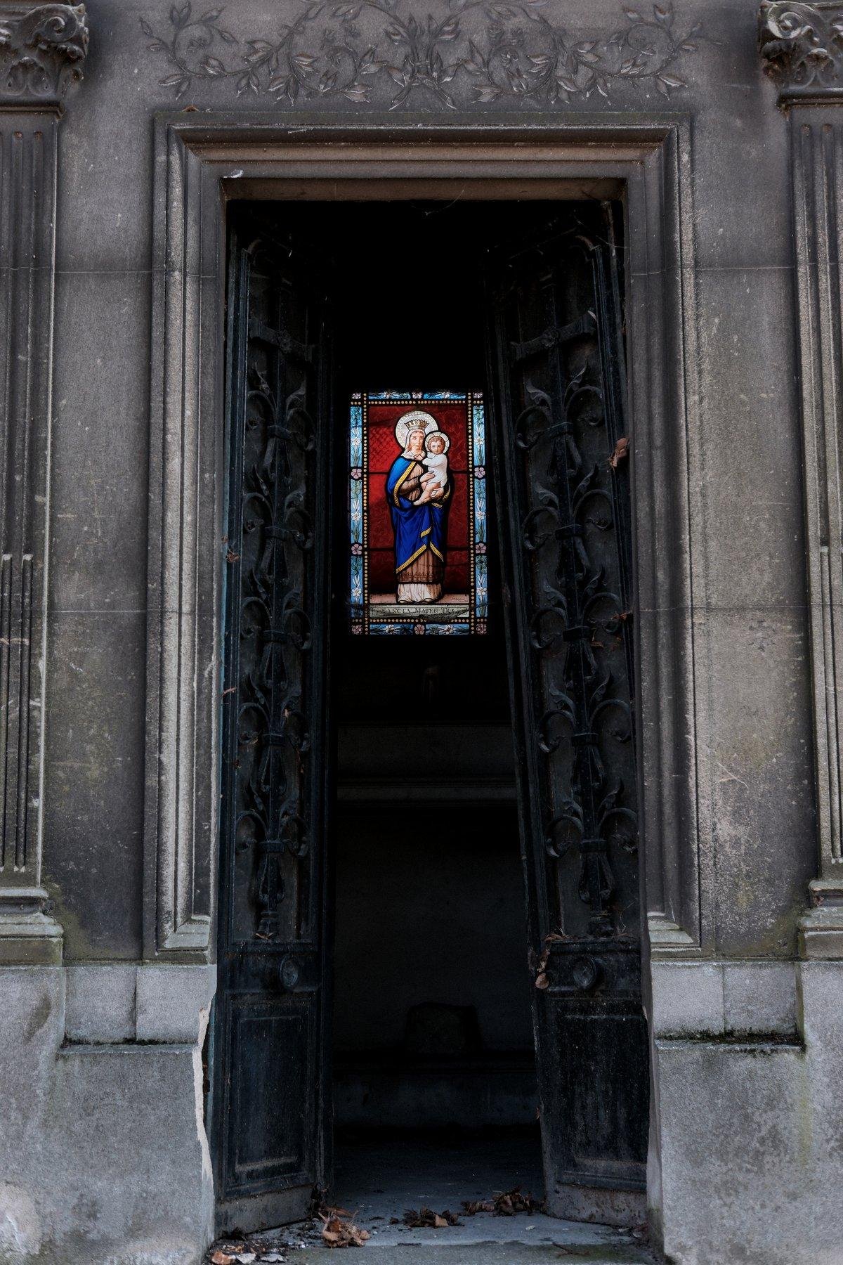 Photo verticale. Un petit vitrail de la vierge Maris et Jesus dans ses bra, sur le mur du fond d’un mausaulée dont les 2 portes sont restées ouvertes