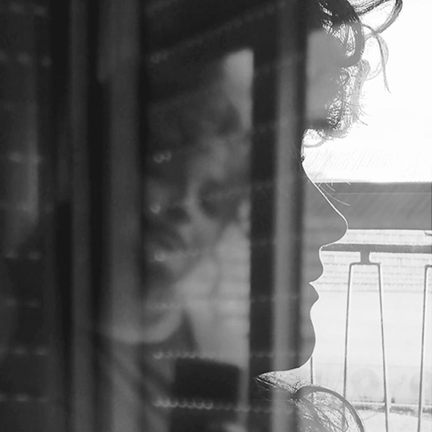 Fotomontaje en blanco y negro en el que se ve una silueta de perfil negra con fondo blanco de una ventana de fondo. En la silueta negra se funde otra foto en la que una cara de una mujer aparece entre reflejos de una ventana. La foto es una fusión de dos fotos, una de ellas que ya de por sí fusiona reflejos