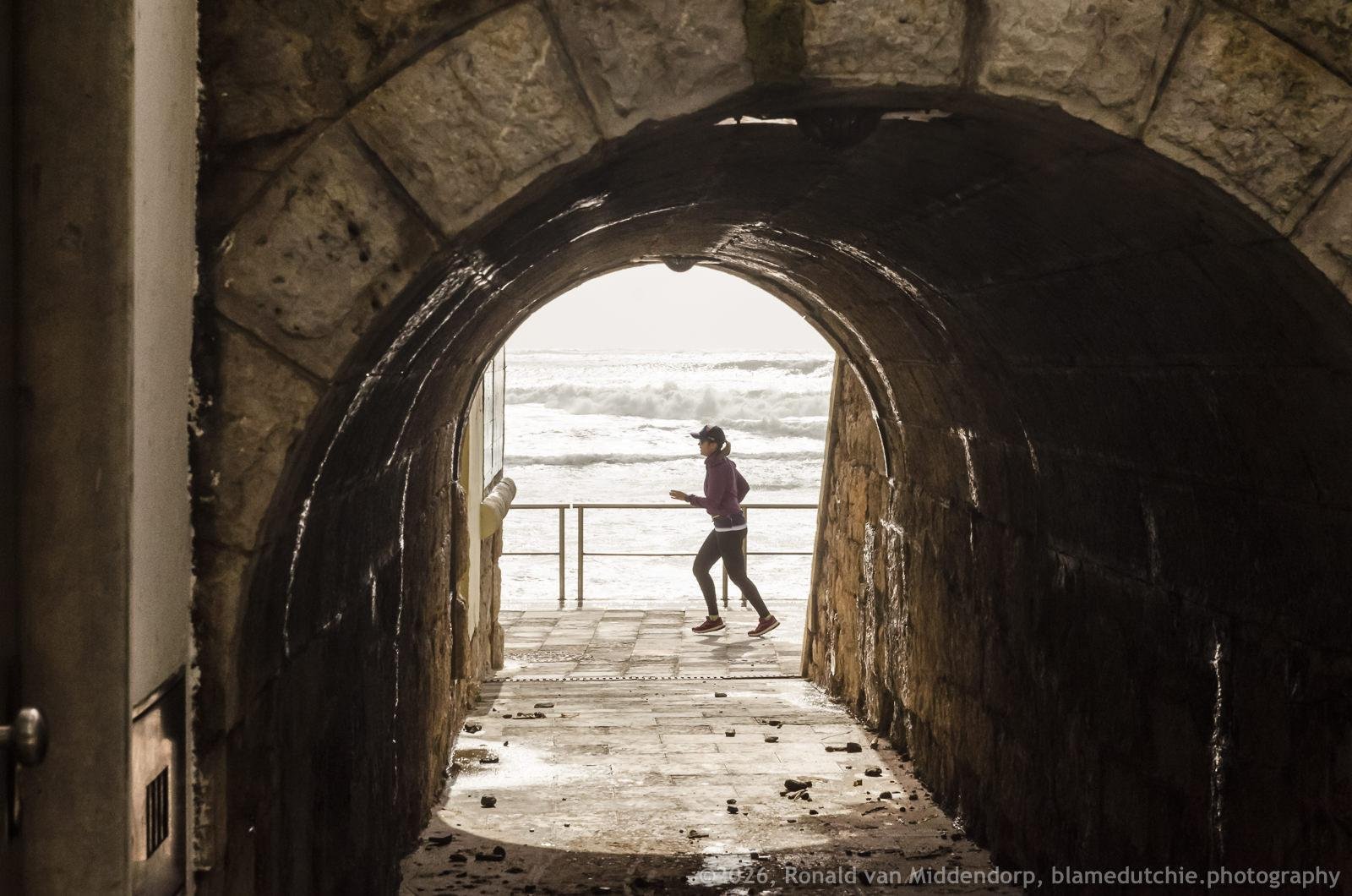 Stone tunnel with wet, dark walls framing a view of the stormy ocean; a jogger in a cap and purple top runs past a railing on a paved walkway at the tunnel’s exit.