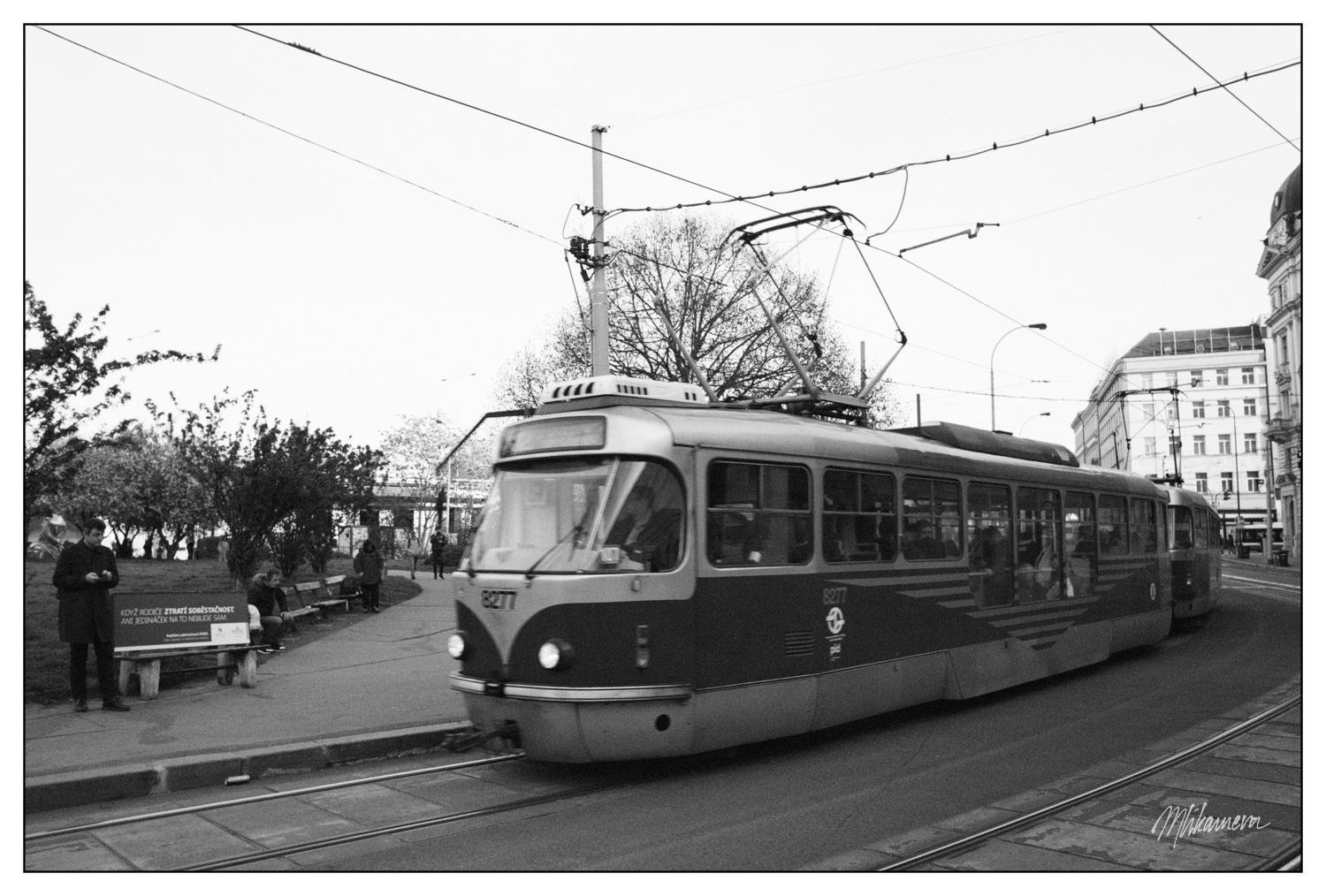 Vintage tram in motion on city tracks, surrounded by urban scenery. Overhead power lines stretch across the sky. A passenger is visible inside, and a person stands near a bench on the sidewalk. Buildings and bare trees are in the background, captured in black and white.