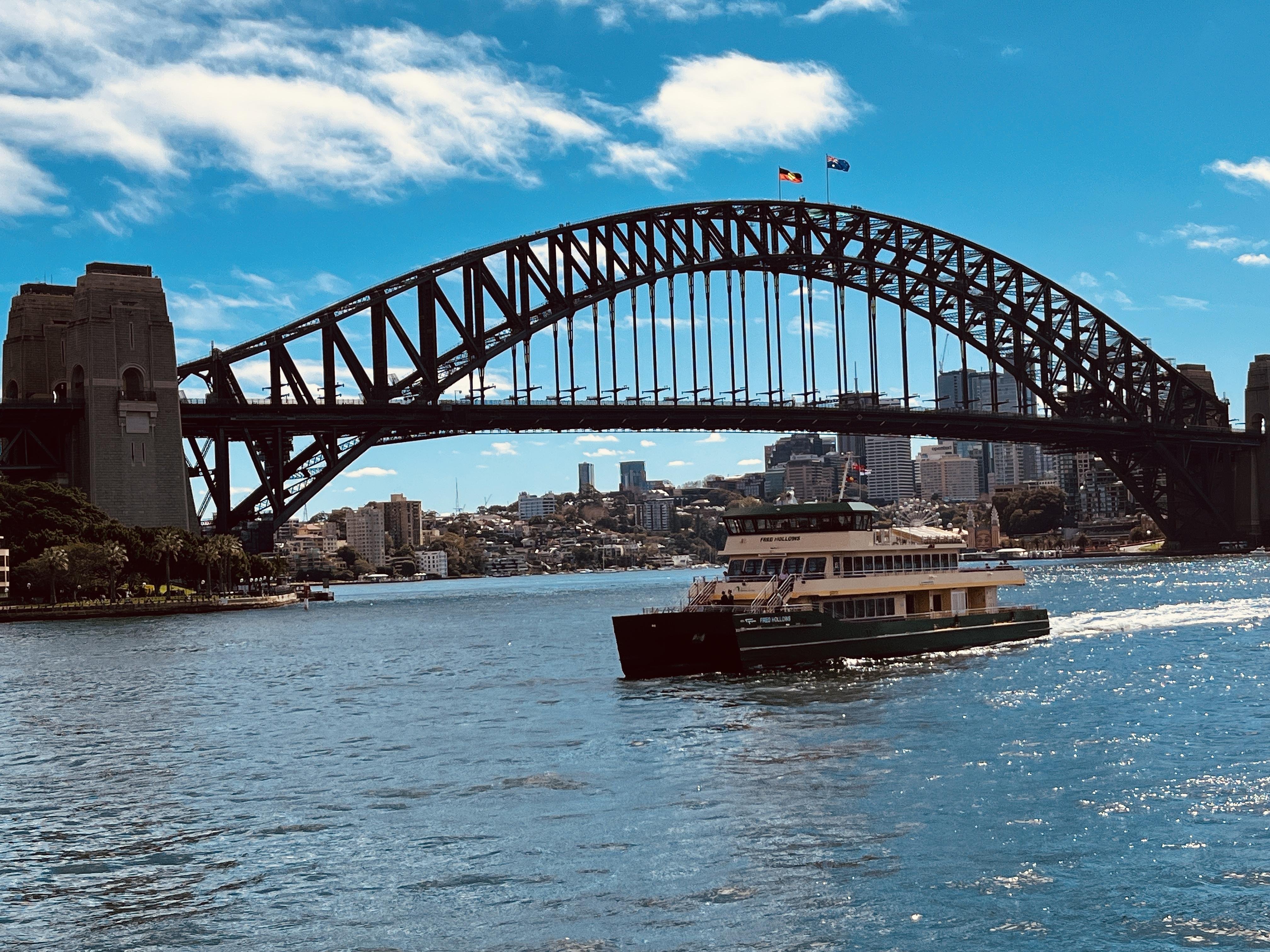 Sydney Harbour Bridge spans across the water under a clear blue sky. A ferry travels on the water in the foreground, with the city's skyline visible in the background.